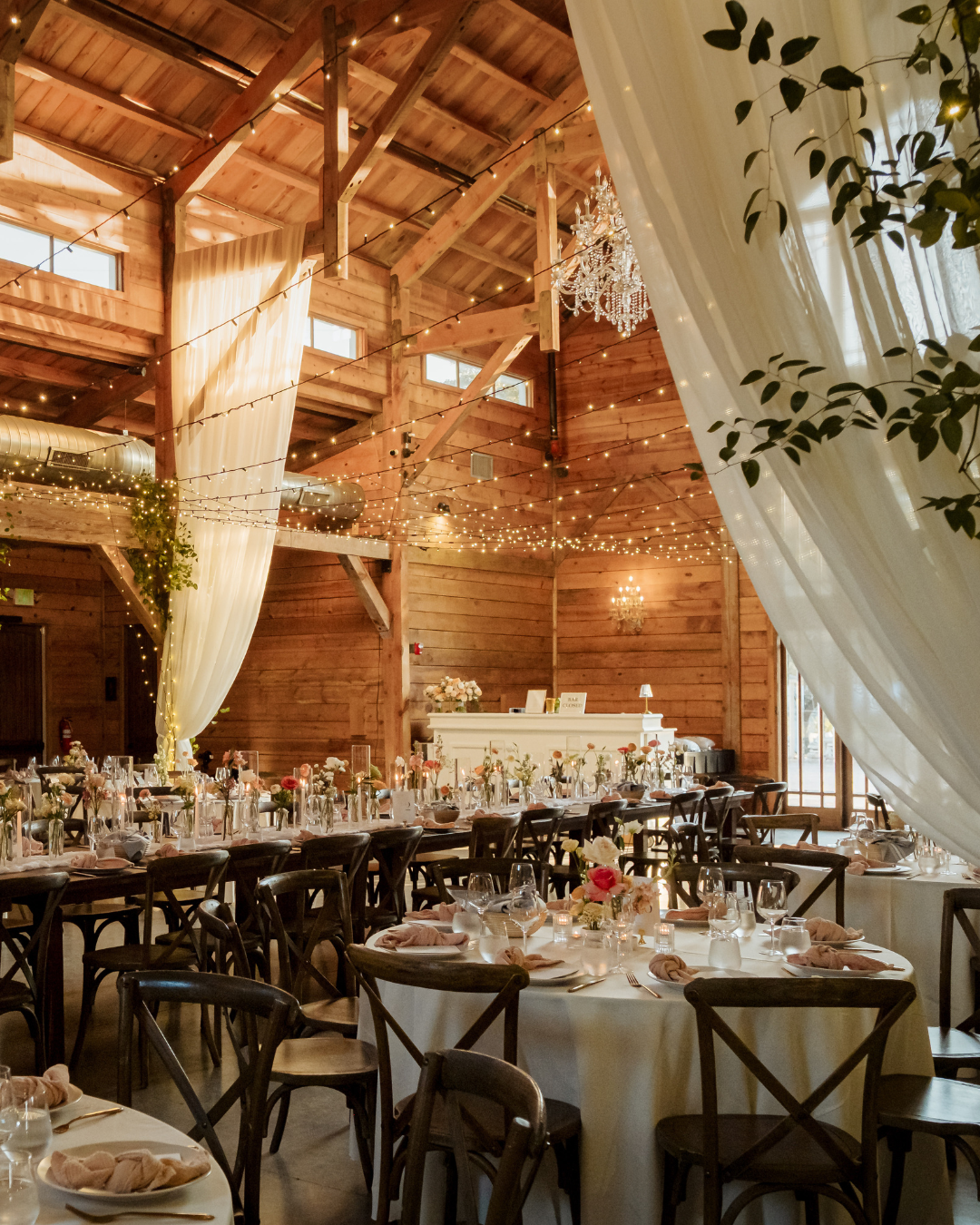 Rustic wooden wedding reception hall with candlelit tables, hanging drapes, and warm string lights