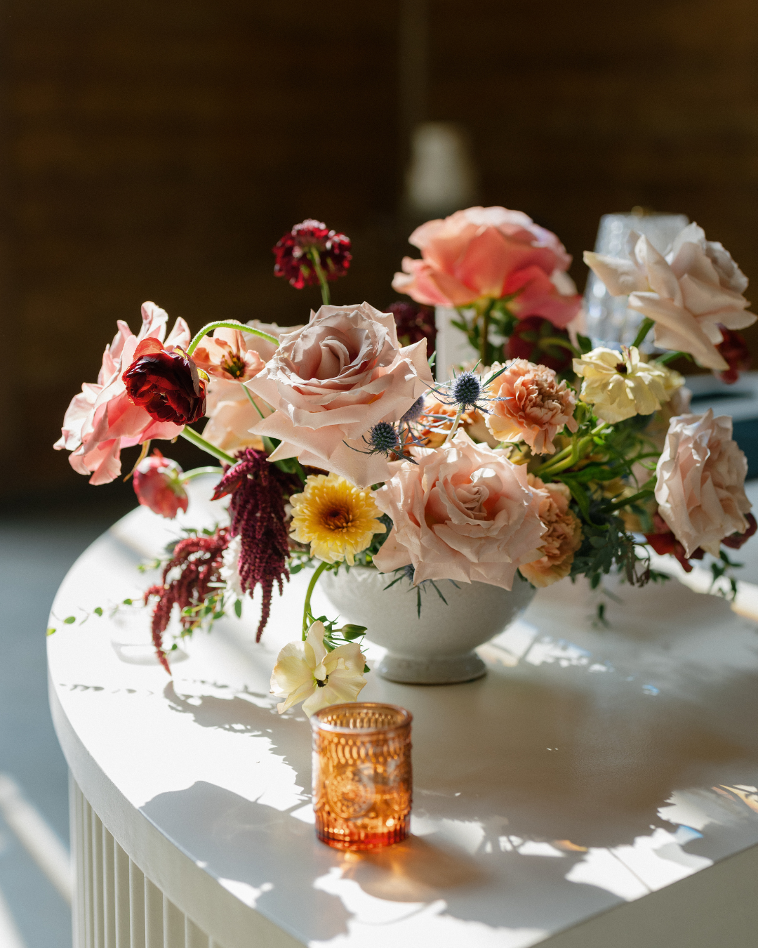 Colorful floral bouquet in a white vase on a sunlit round table with a small amber glass candleholder