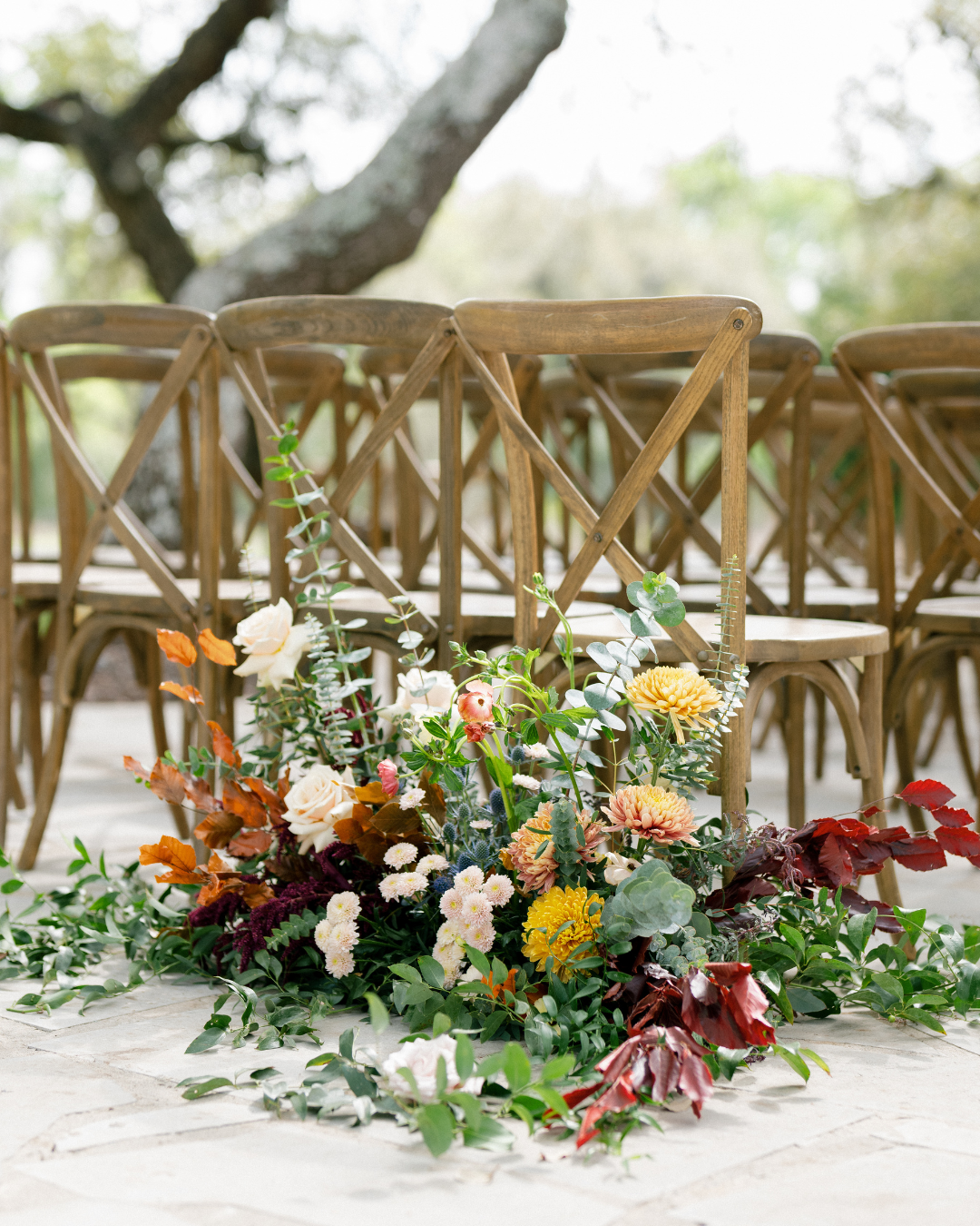 Floral arrangement with white, orange, yellow, and burgundy flowers in front of wooden chairs outdoors