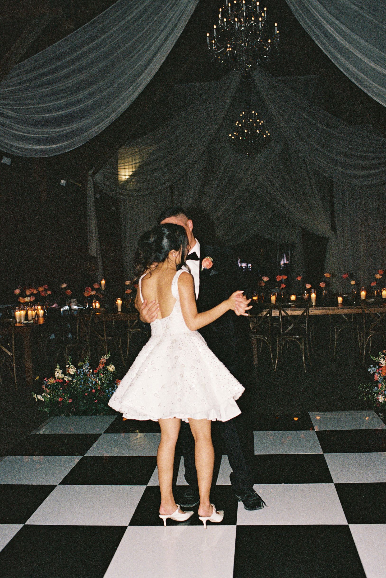 A couple dances on a black-and-white checkered floor in a dimly lit, elegantly decorated venue with draped fabric.