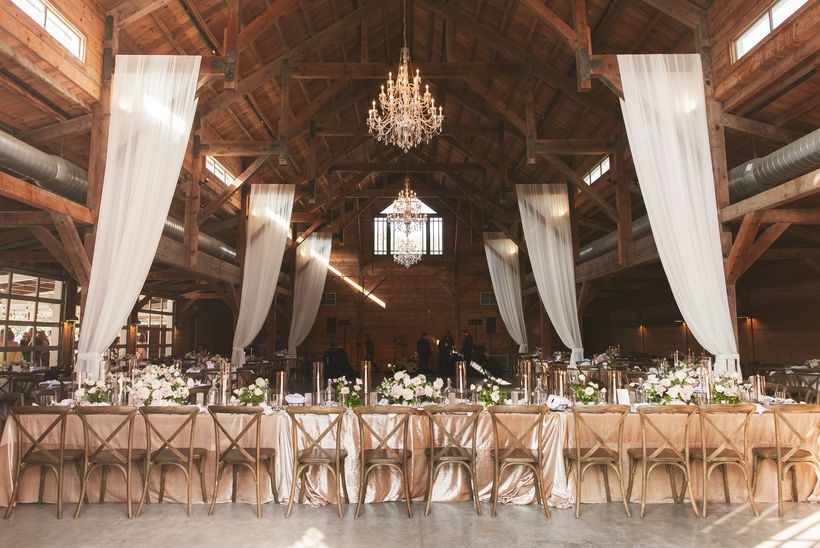 Elegant banquet hall with long floral tables, chandeliers, and white drapes under a wooden ceiling