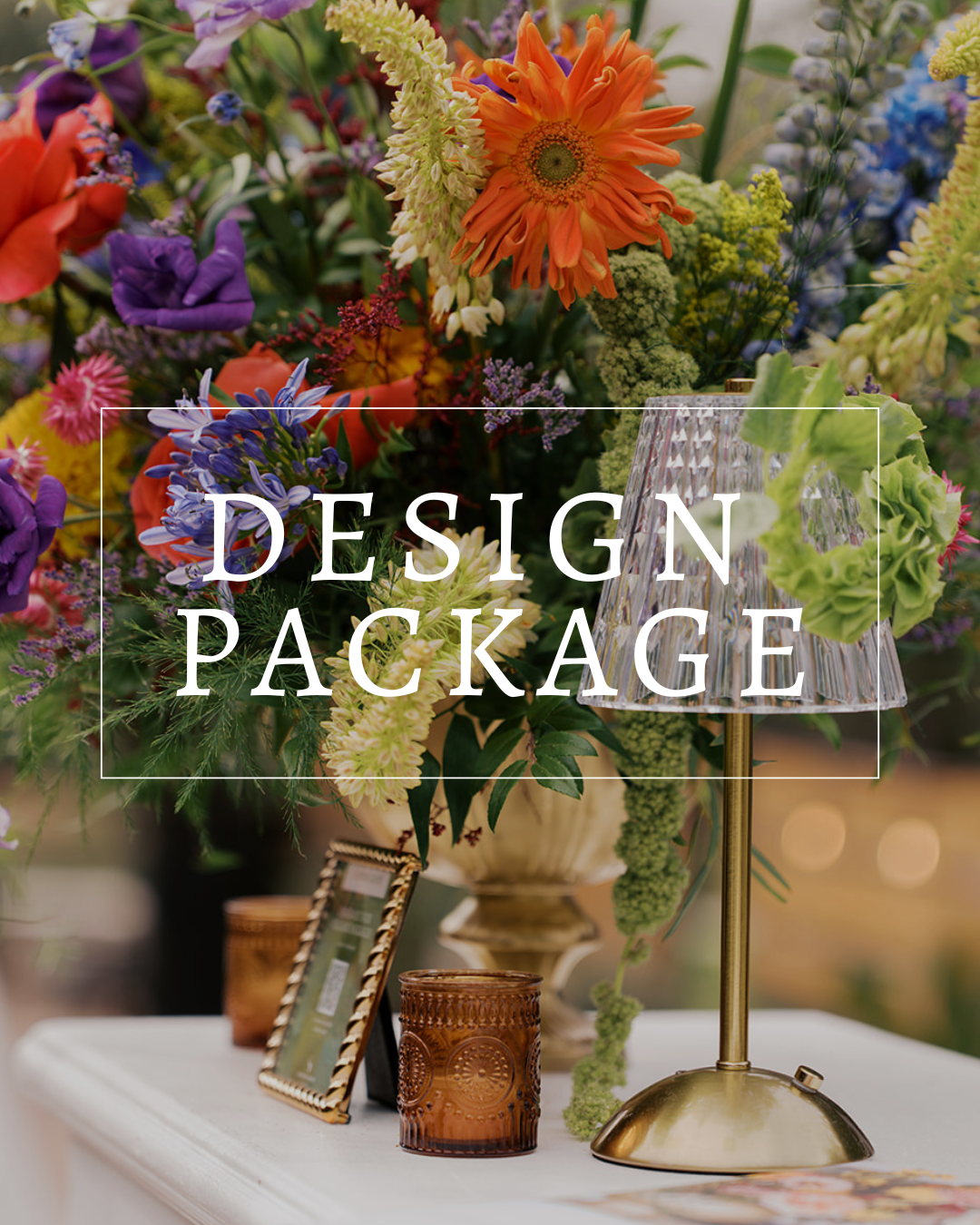 A vibrant, multi-colored floral arrangement sits on a table next to a brass lamp and a small candle in a glass holder.