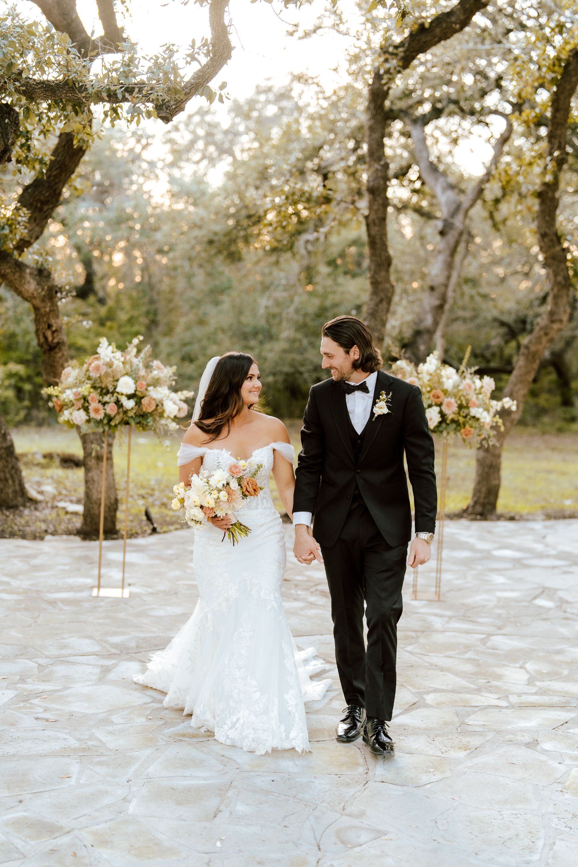 A couple in wedding attire walks hand-in-hand on a stone patio surrounded by trees and floral arrangements.