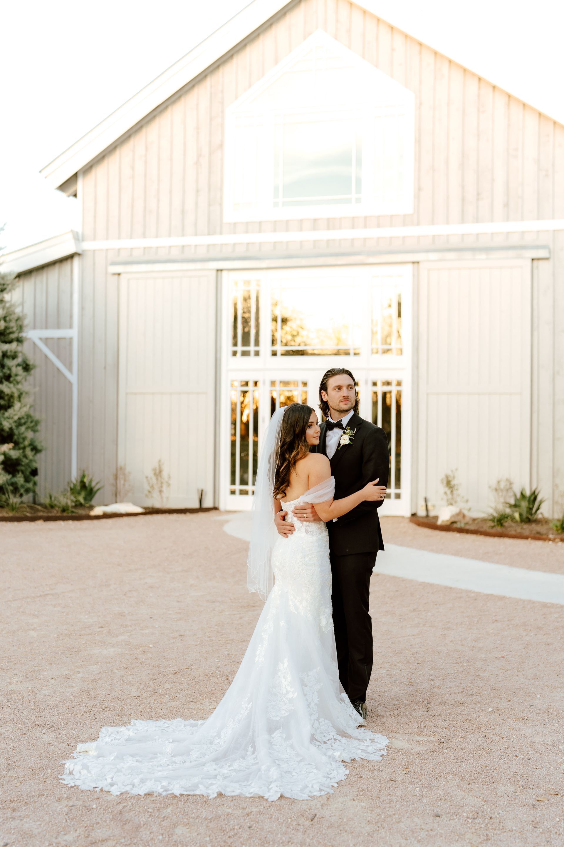 A bride in a white lace gown and a groom in a black suit pose together in front of a light-colored barn at sunset at The Addison Grove.
