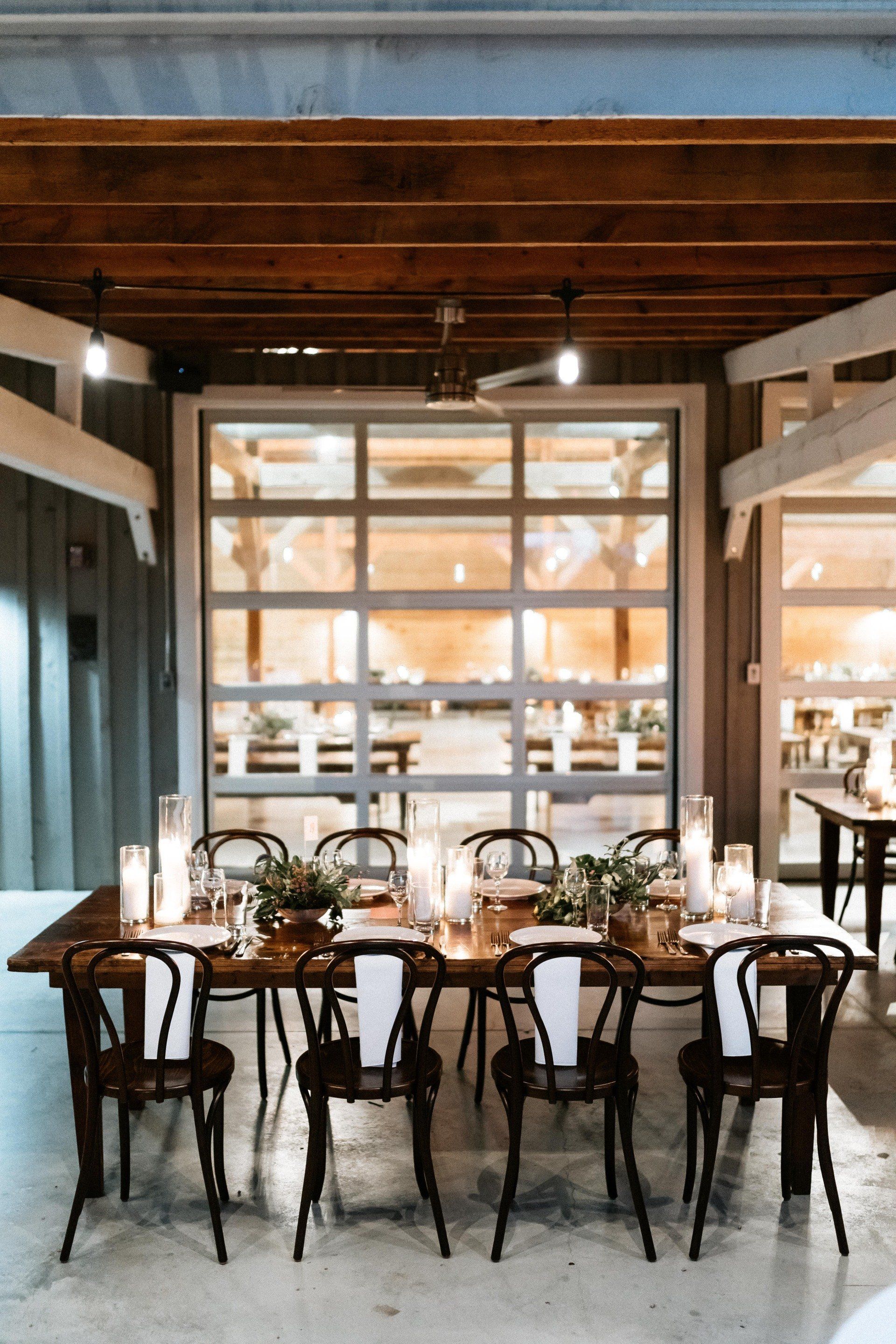 A set dining table with candles and chairs in a rustic, open-air venue with a large glass garage door in the background.