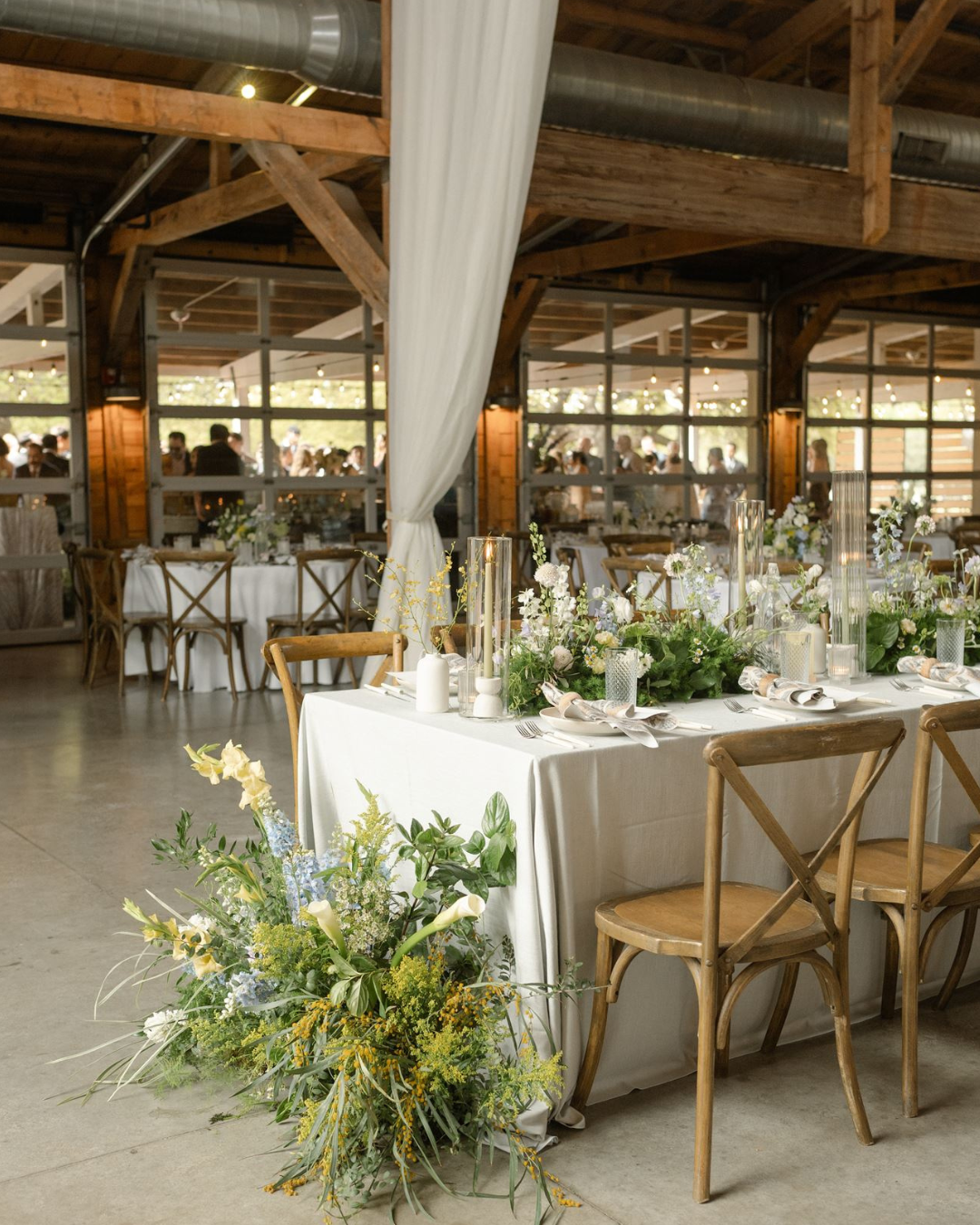 Rustic wedding reception table with white linens, floral greenery, and wooden chairs in a barn venue