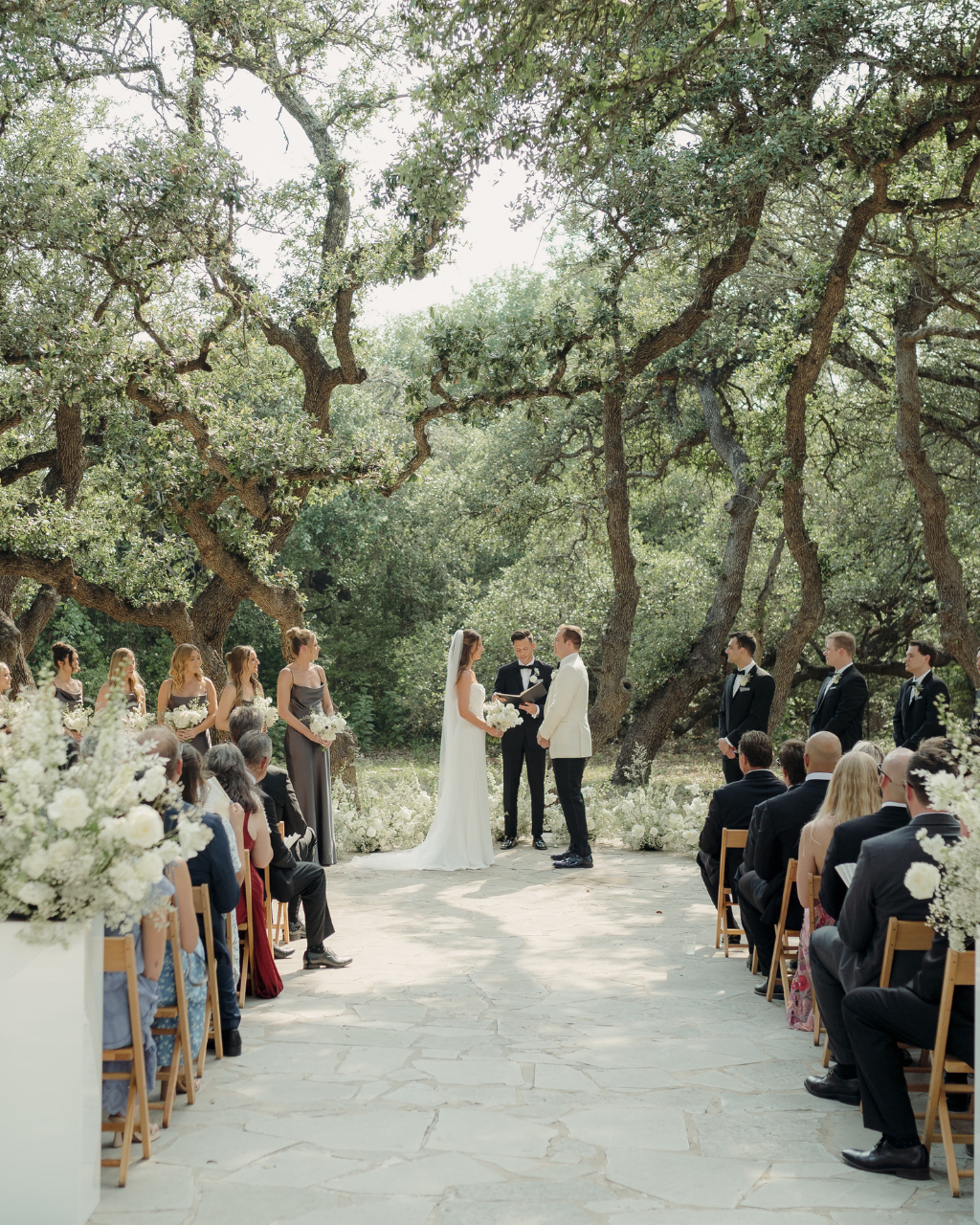 Outdoor wedding ceremony under trees with guests seated along a stone aisle, bride and groom at the altar