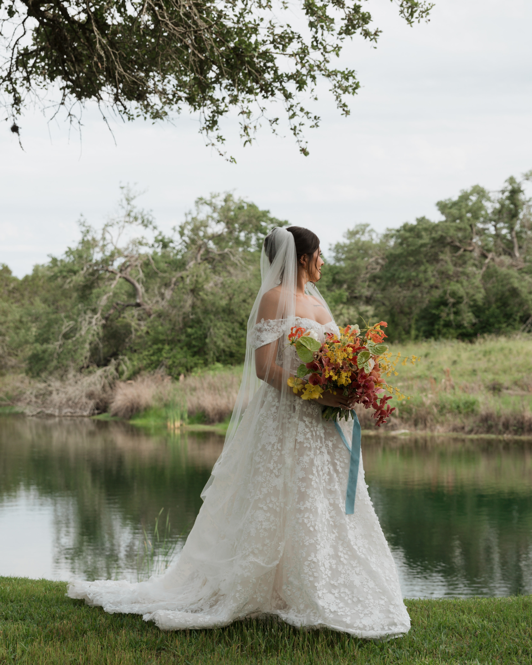 A bride in a lace off-the-shoulder gown and veil holds a vibrant fall bouquet by a river with trees in the background.