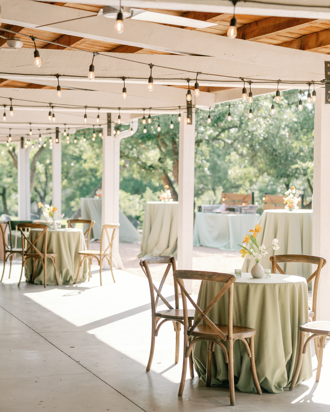 Open-air event patio with round tables, green tablecloths, and string lights under a wooden pergola.