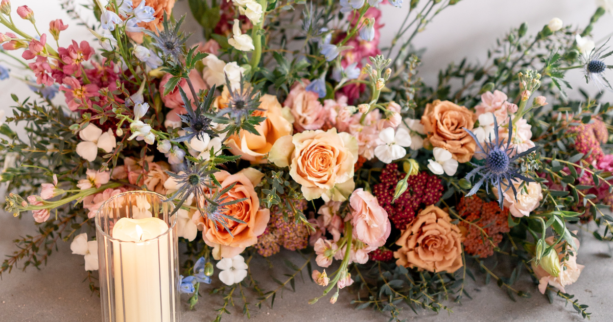 Pastel floral arrangement with peach roses, pink blooms, and greenery on a table beside a candle