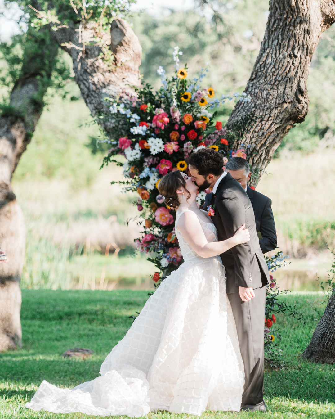 A bride and groom kiss outdoors beneath a large floral arrangement nestled in the crook of two trees.