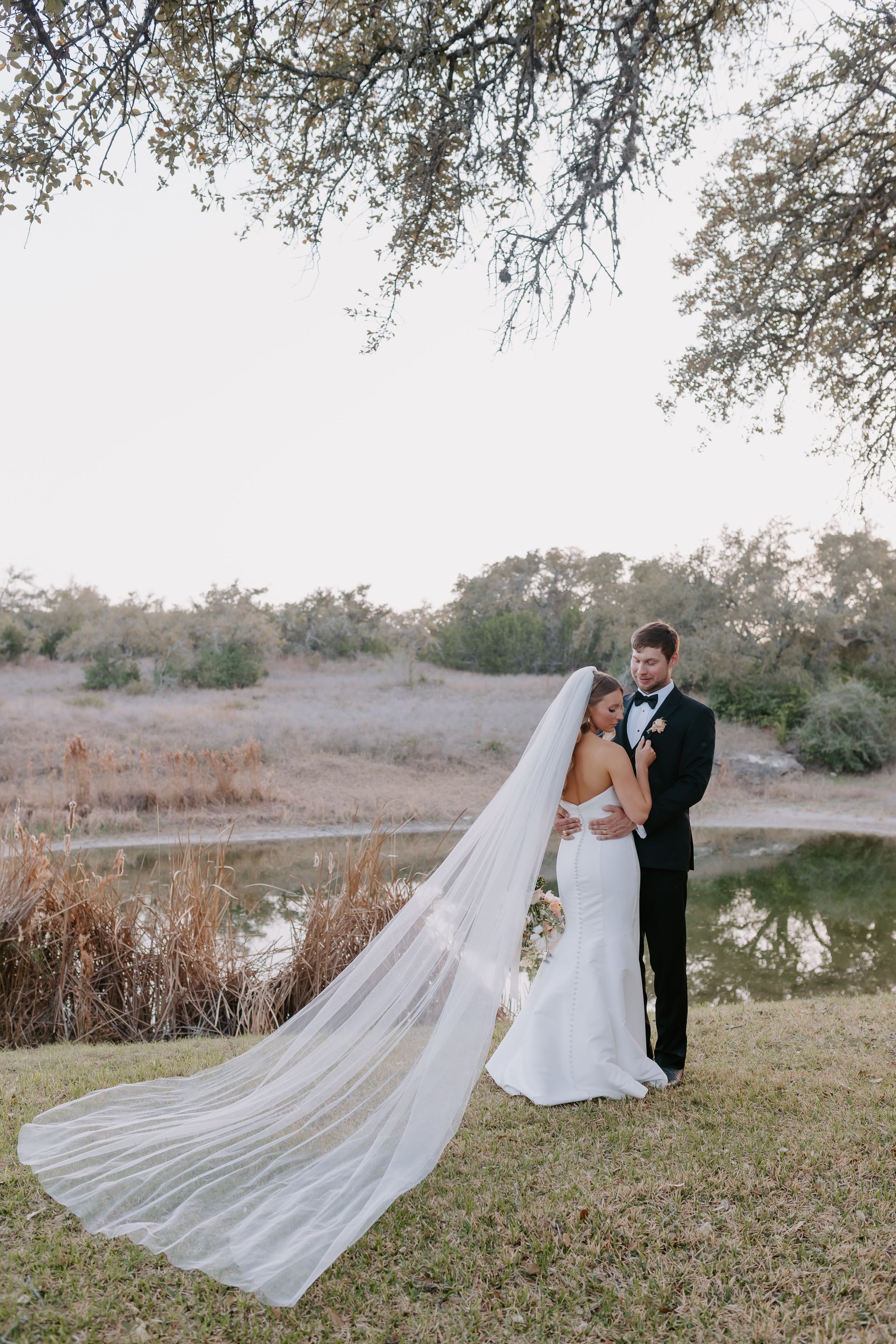 A wedding couple embraces by a pond, with a long, flowing veil trailing across the grass beneath trees.