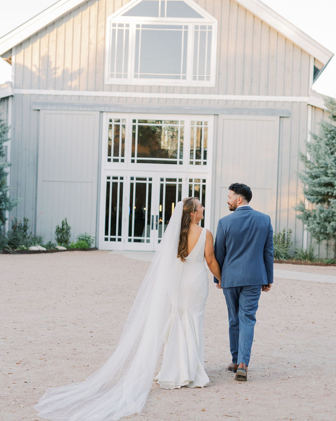 Bride and groom walking toward a white barn venue, bride in a long veil and train