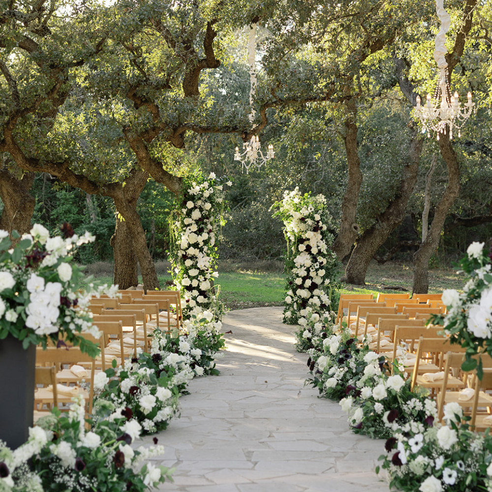 Outdoor wedding aisle with white floral arrangements and gold chairs under trees