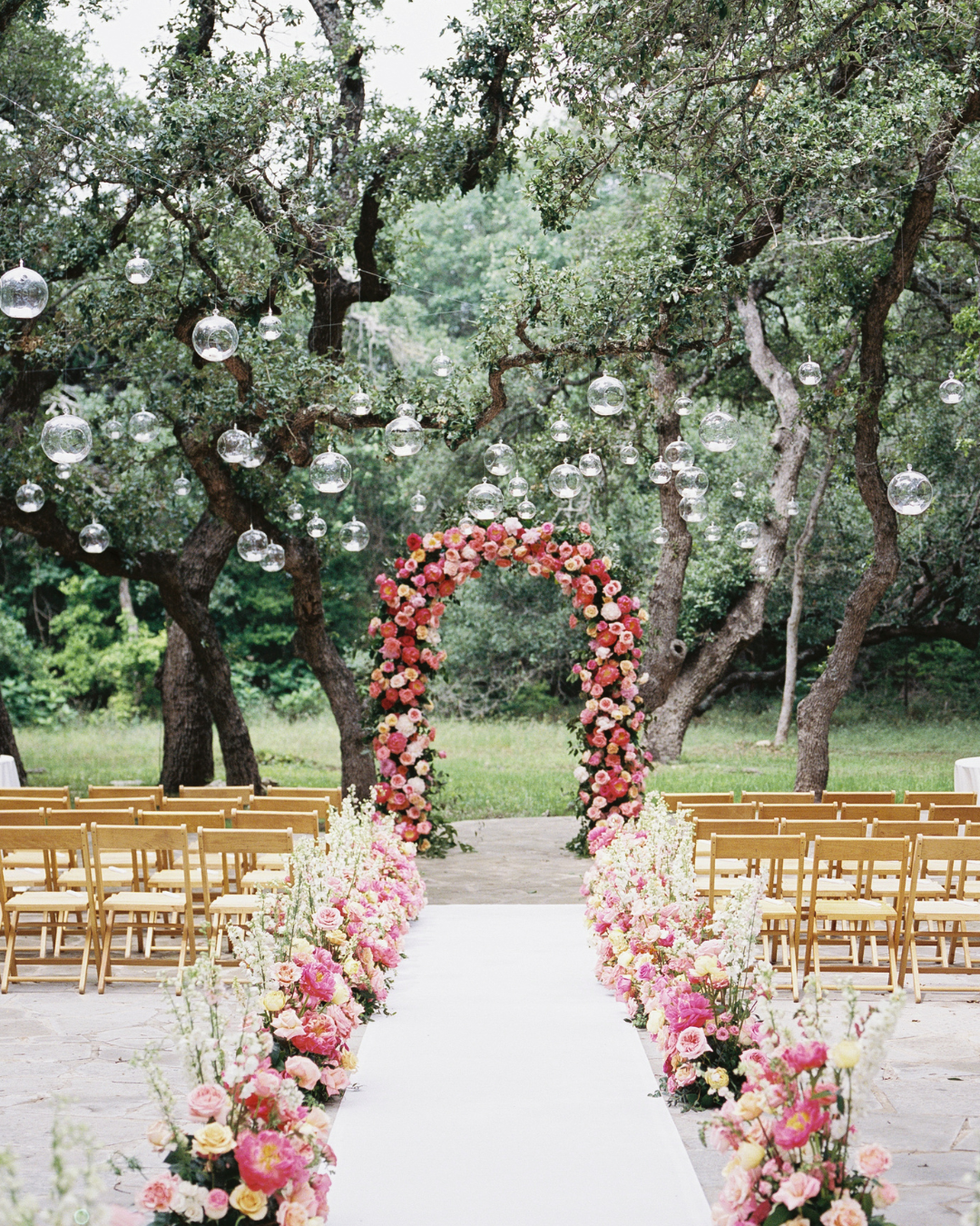 An outdoor wedding ceremony setup featuring a floral arch, a white aisle, and rows of wooden chairs under trees.