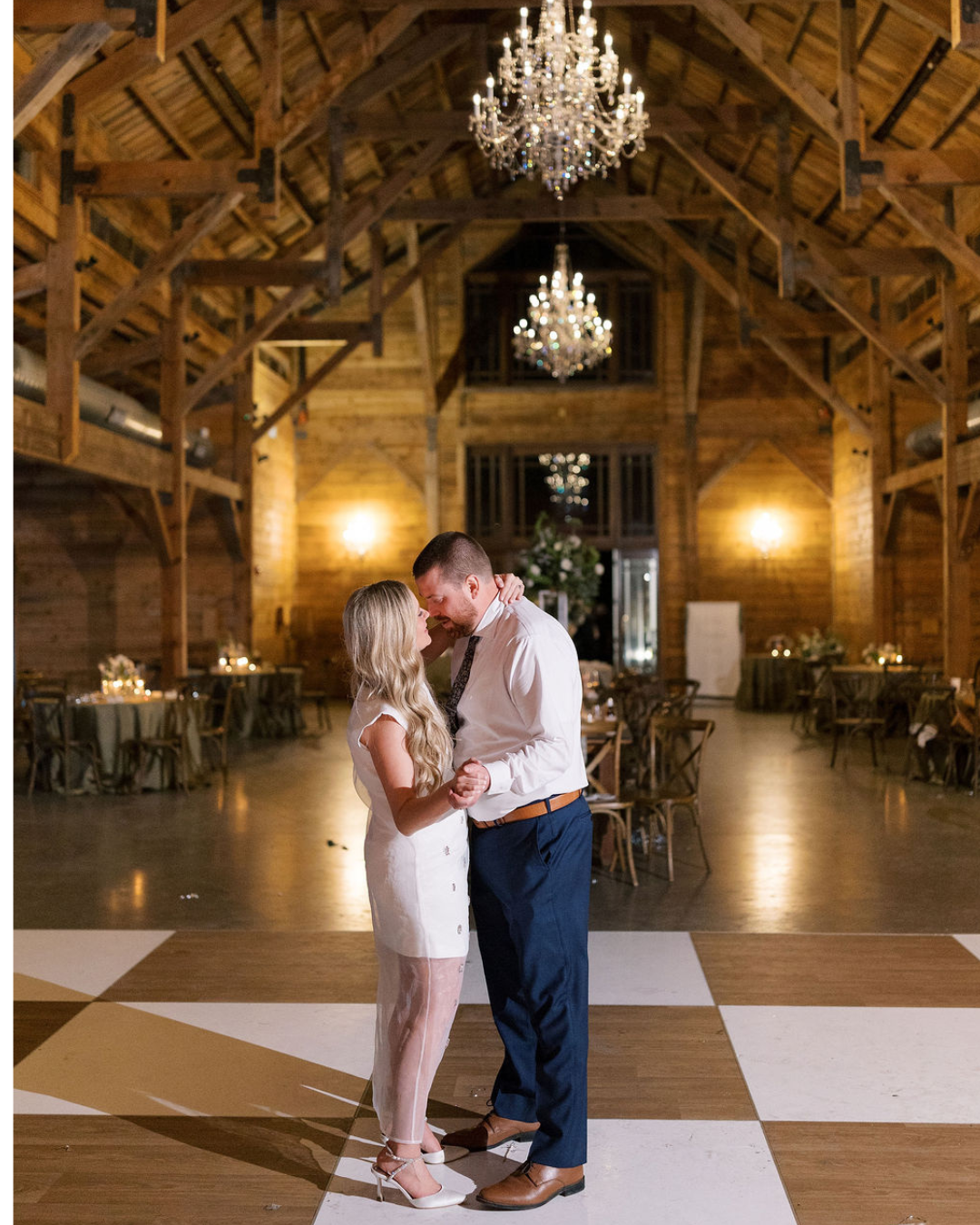 A couple dances together on a checkered floor in a rustic, chandelier-lit barn.