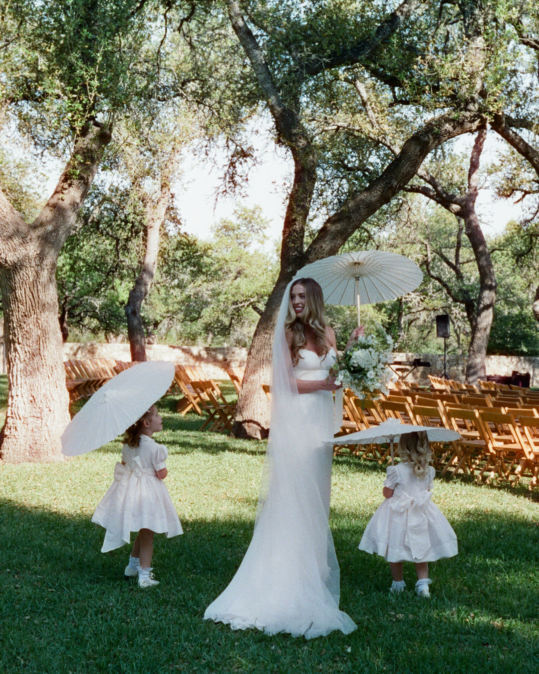 A bride in a white wedding gown holding a parasol walks outside between two flower girls with parasols under large trees.