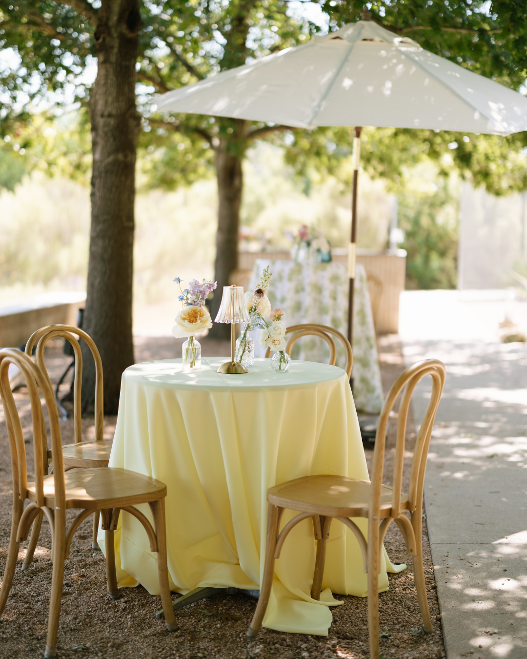 A small, round table with a light yellow tablecloth and wooden chairs set outdoors under a white patio umbrella.