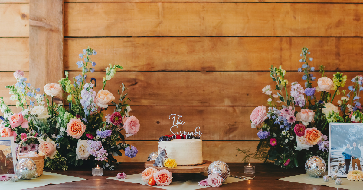 Floral wedding dessert table with pastel bouquets, a small cake, and decorative treats against a wooden wall