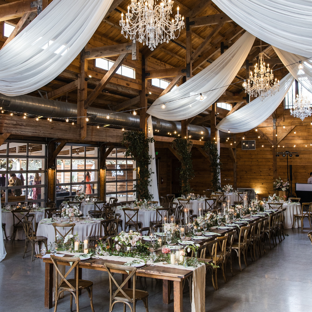 Rustic barn wedding reception with long tables, white drapes, chandeliers, and warm wood beams