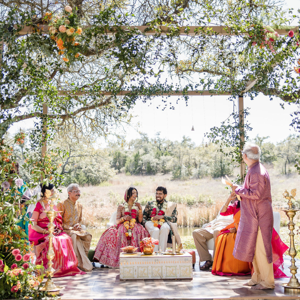 People seated at a colorful outdoor gathering under a decorated canopy, with a standing speaker nearby.