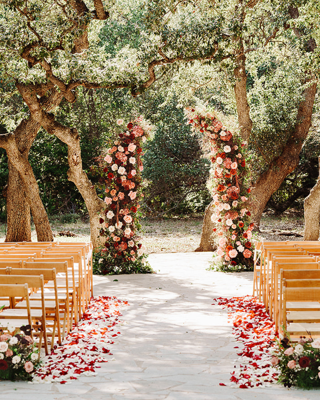 Outdoor wedding aisle with wooden chairs, floral arches, and rose petals under trees