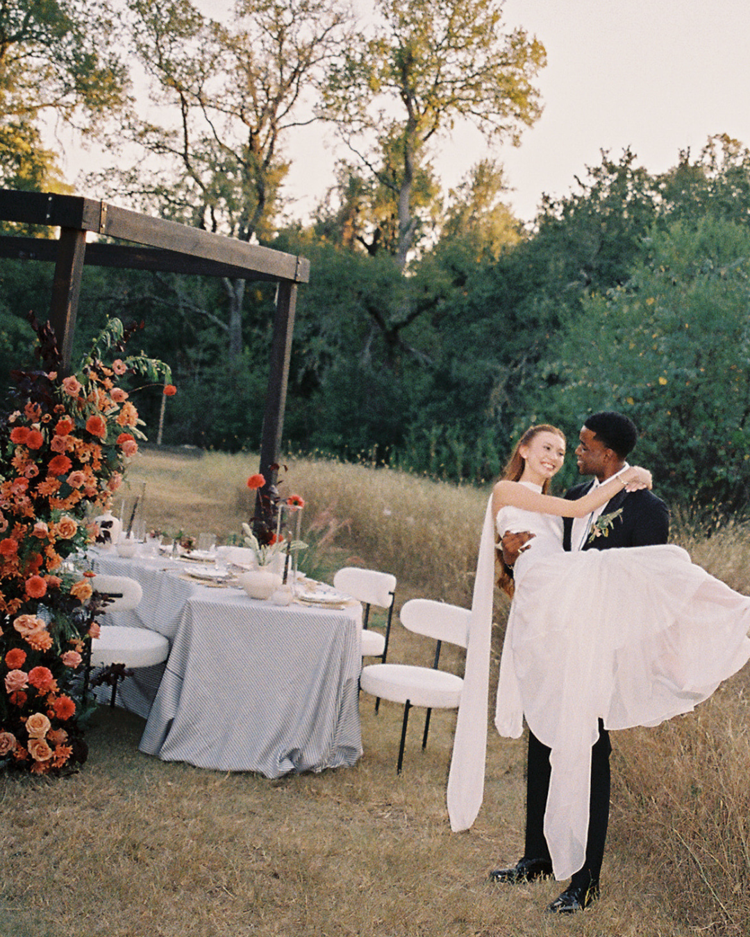 Couple in wedding attire dancing outdoors beside a floral-decorated table at sunset