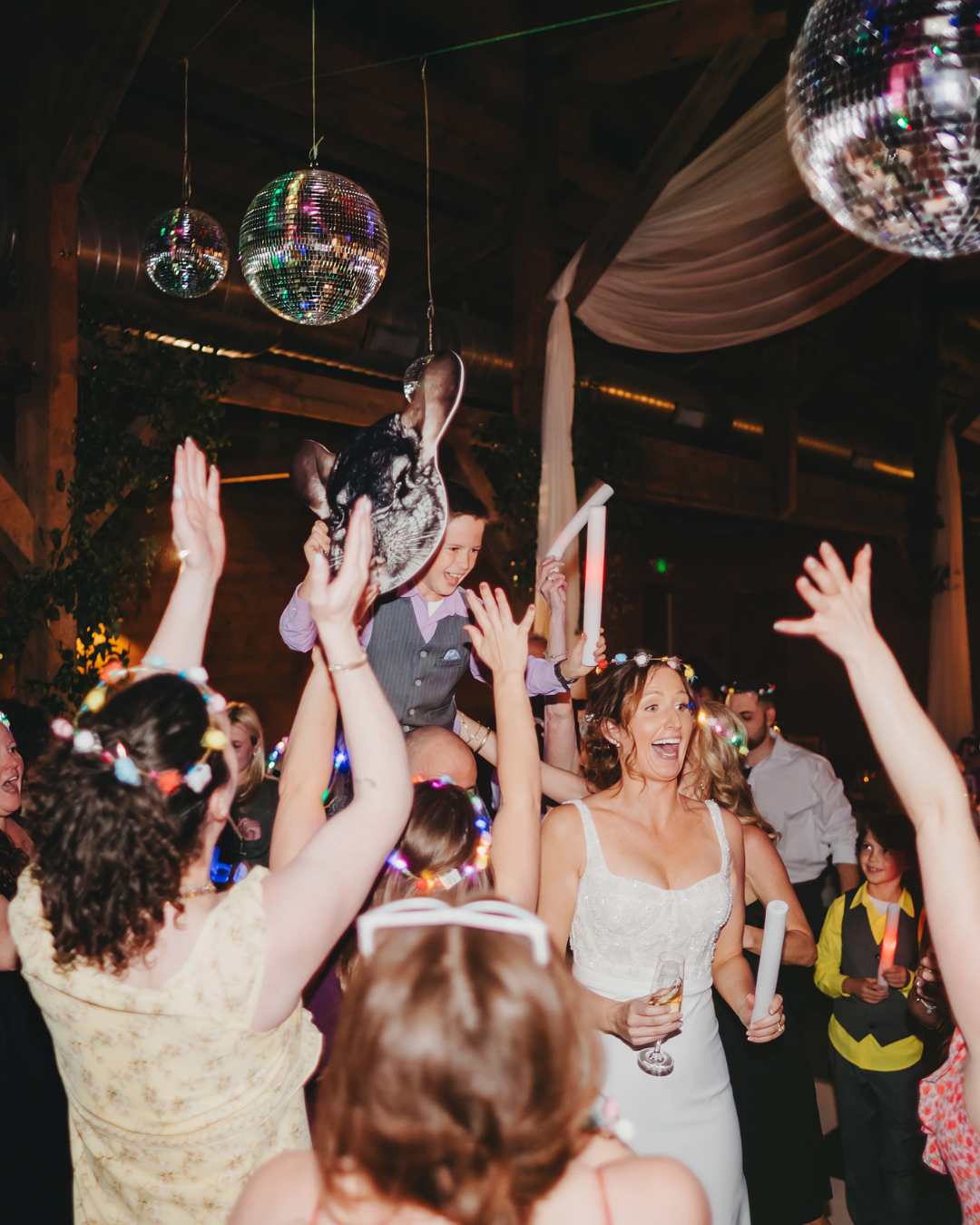 A wedding reception scene with guests dancing beneath disco balls while someone is lifted into the air.