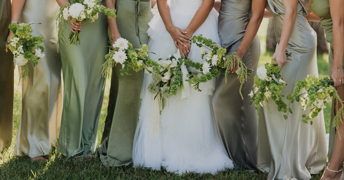 Bridesmaids in pastel dresses hold white-flower bouquets beside a bride in a white gown outdoors.