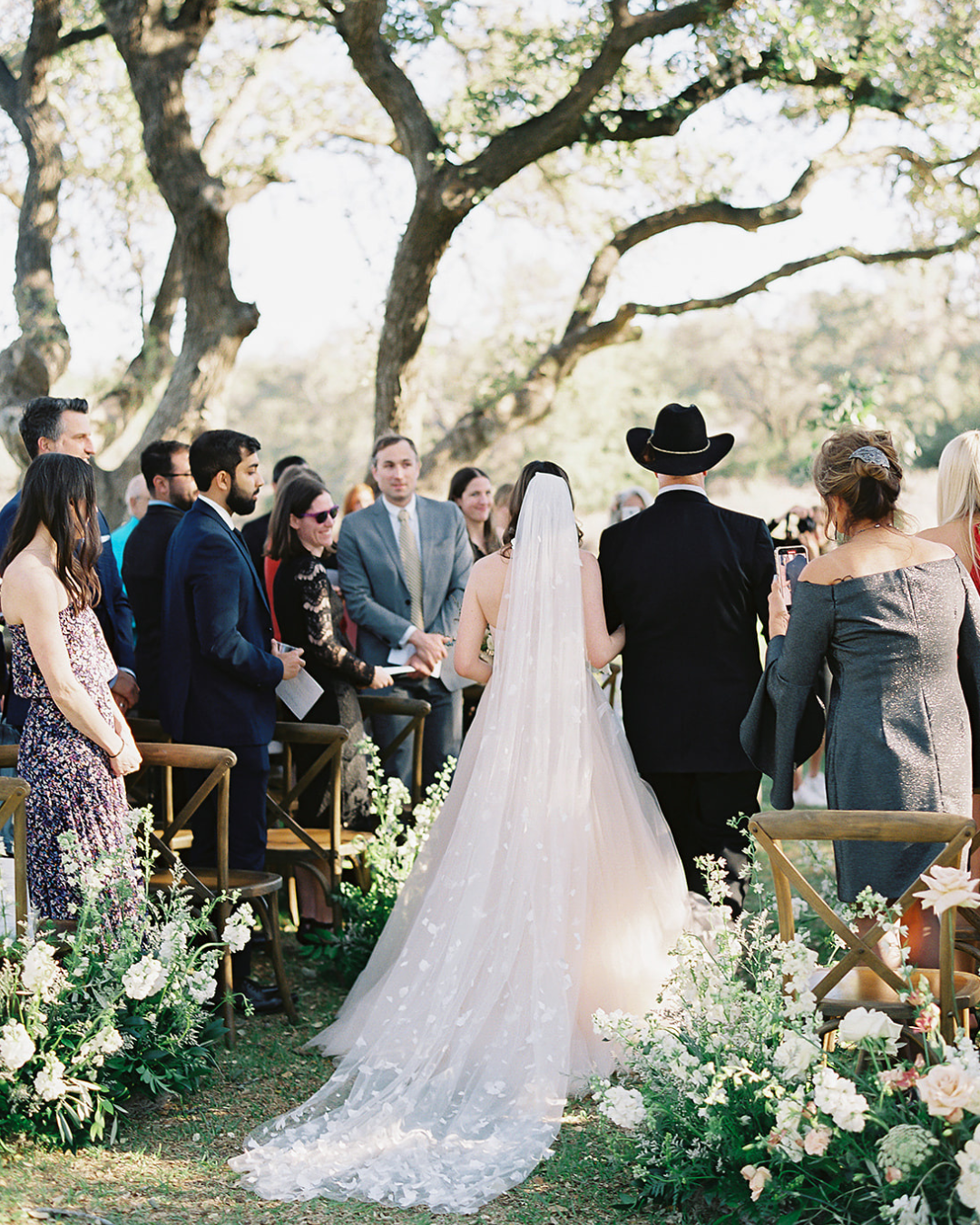 Bride walks down an outdoor aisle with guests and officiant under trees at a wedding ceremony
