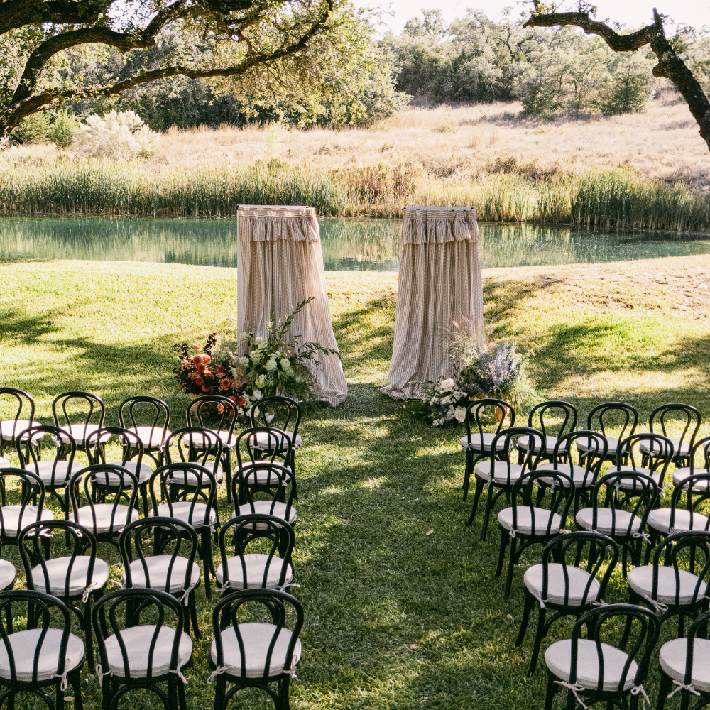 Outdoor wedding ceremony setup with white chairs, decorated aisle, and draped altar by a lakeside lawn