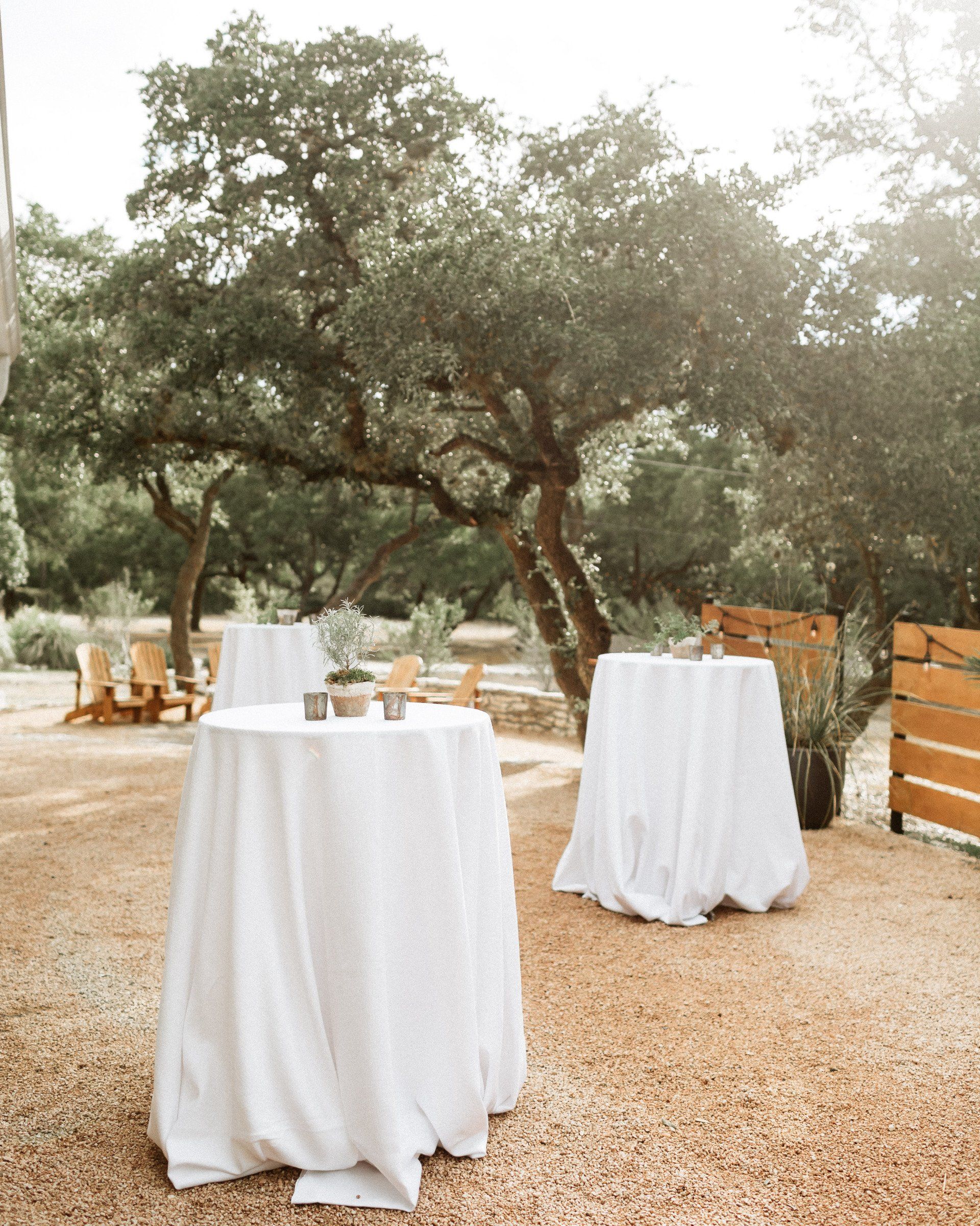 White cocktail tables sit on a gravel patio under large trees, set up for an outdoor event.