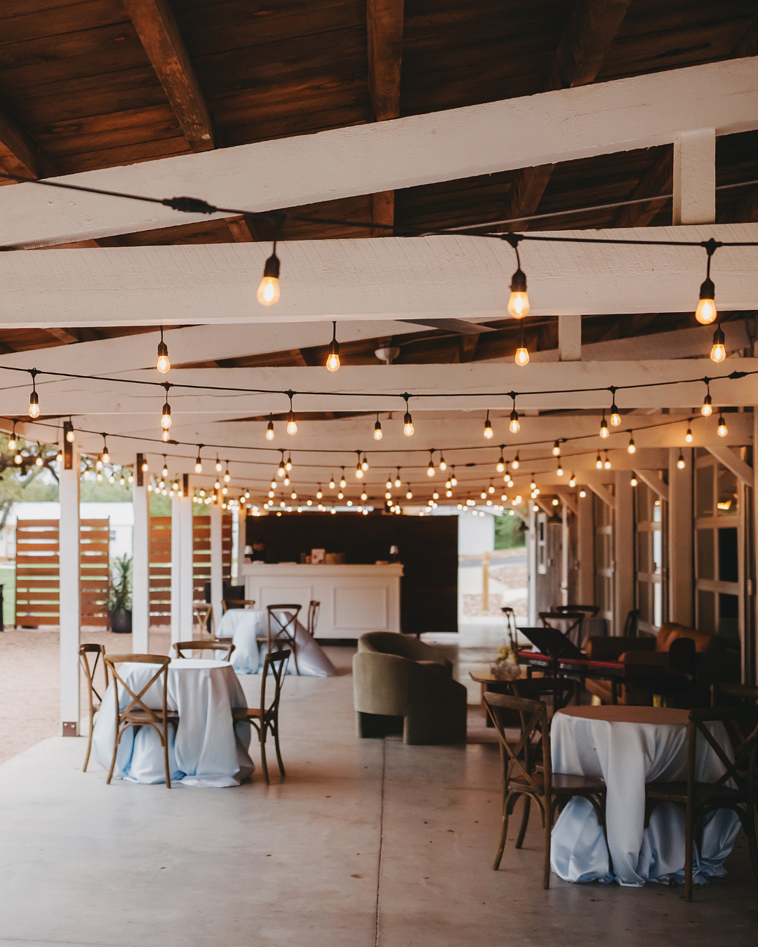 An outdoor event venue with white-clothed tables, wooden chairs, and string lights hanging from wooden rafters.