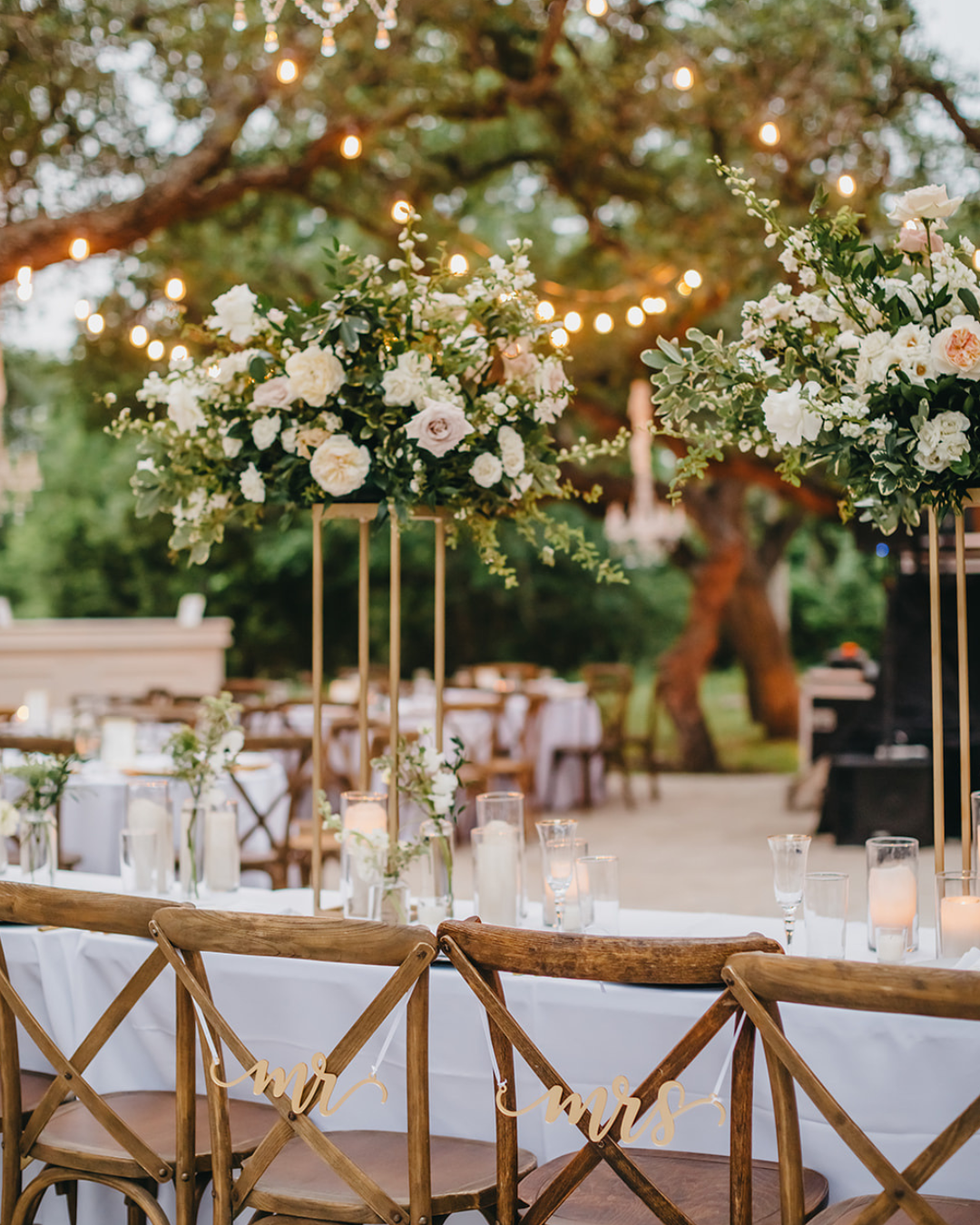 An outdoor wedding reception setup with wooden cross-back chairs, floral centerpieces on gold stands, and string lighting.
