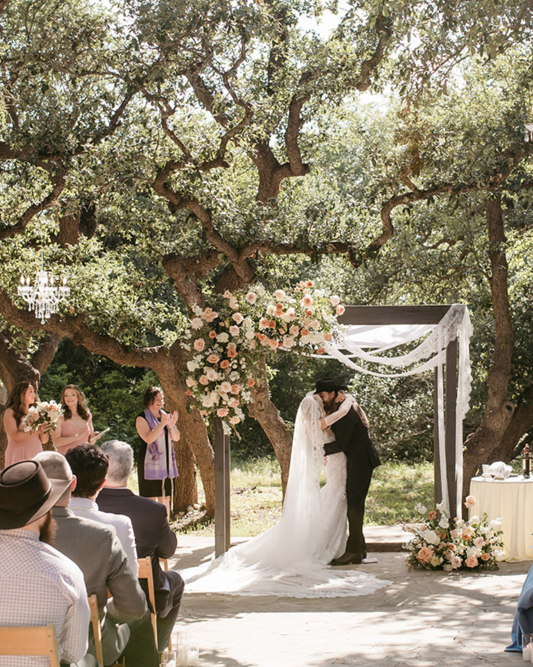 Bride and groom kiss under a floral wedding arch at an outdoor ceremony under trees
