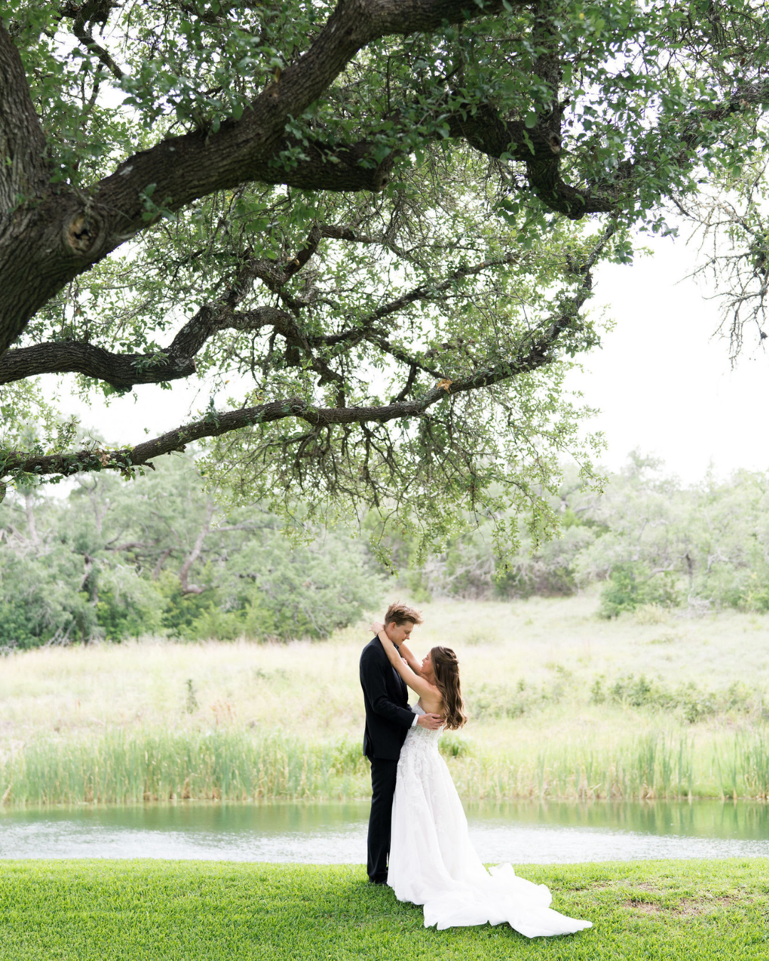 Couple in formal attire embracing by a lakeside under a large tree, with a white gown and black tuxedo.