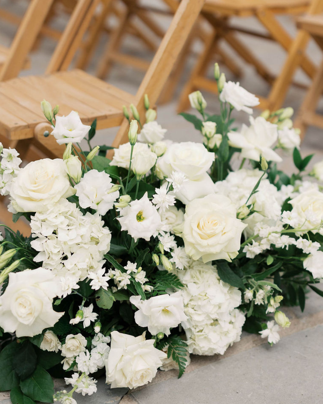 White floral arrangements on the floor beside wooden chairs at an outdoor ceremony setting