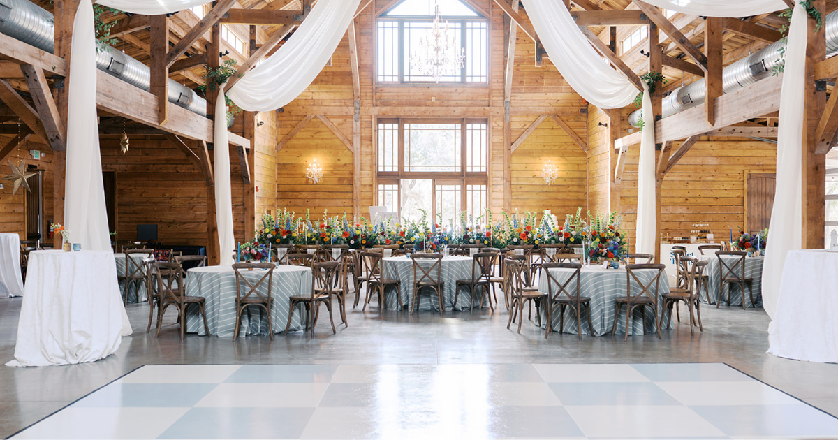 Rustic wedding reception hall with long banquet tables, tall windows, and white draped decor