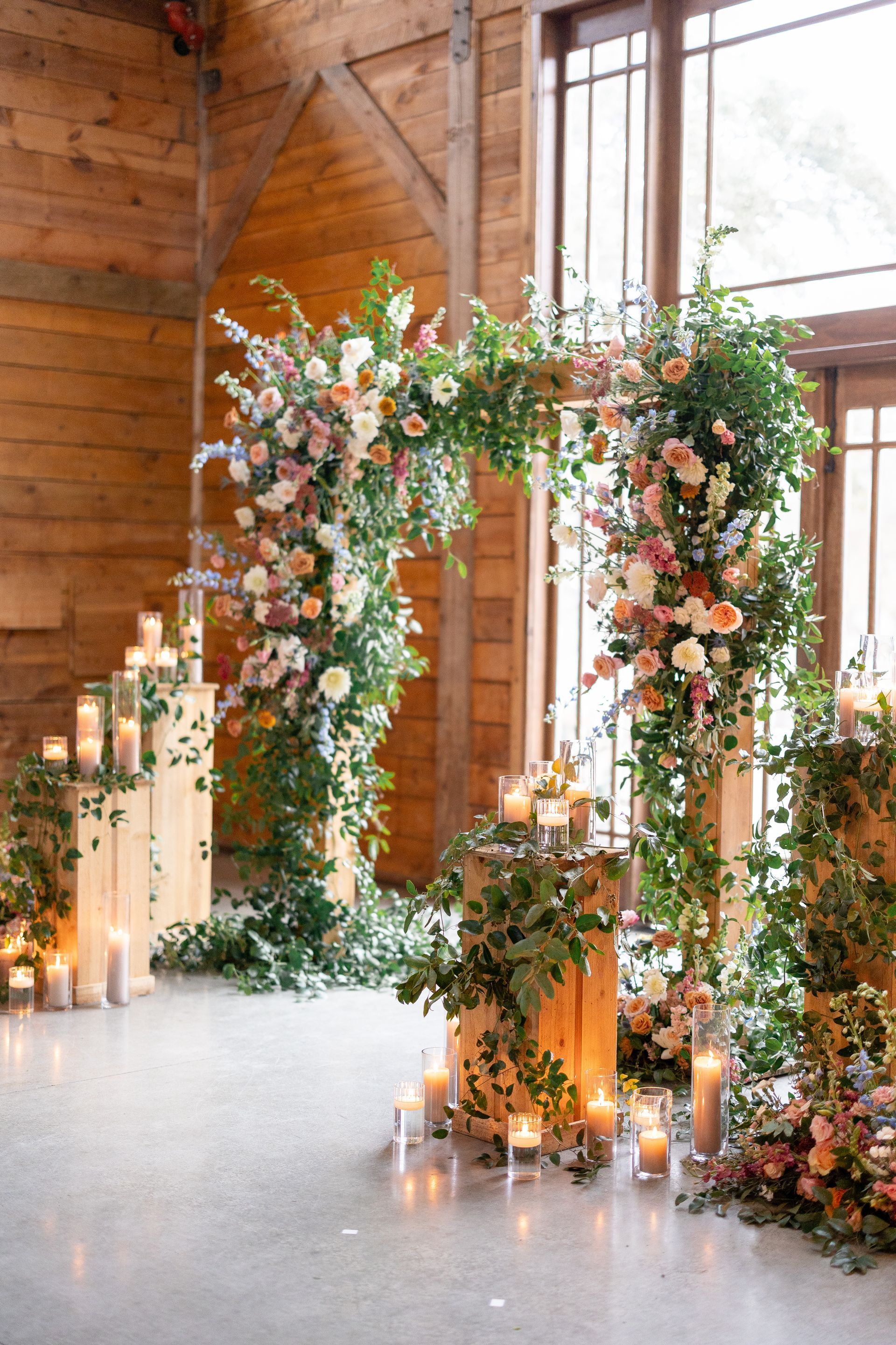 A floral wedding arch stands on wooden pedestals surrounded by numerous lit candles inside a rustic barn.