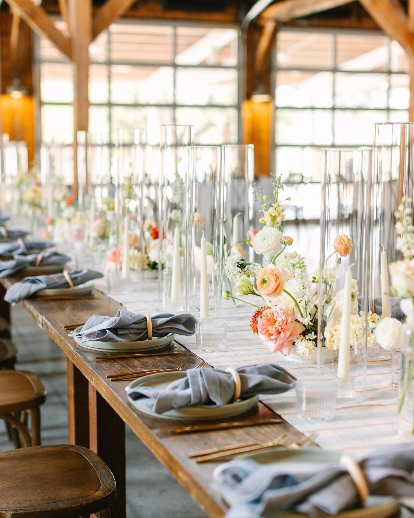 A long wooden banquet table set for a wedding with floral centerpieces, tall glass vases, and blue napkins in wooden rings.