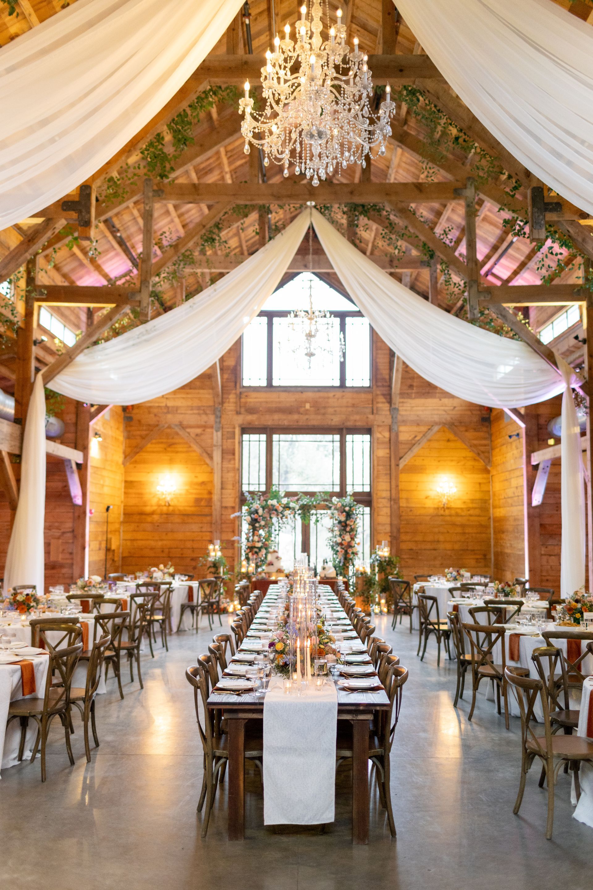 A rustic wooden barn wedding venue featuring a long banquet table, elegant white fabric draping, and a crystal chandelier.