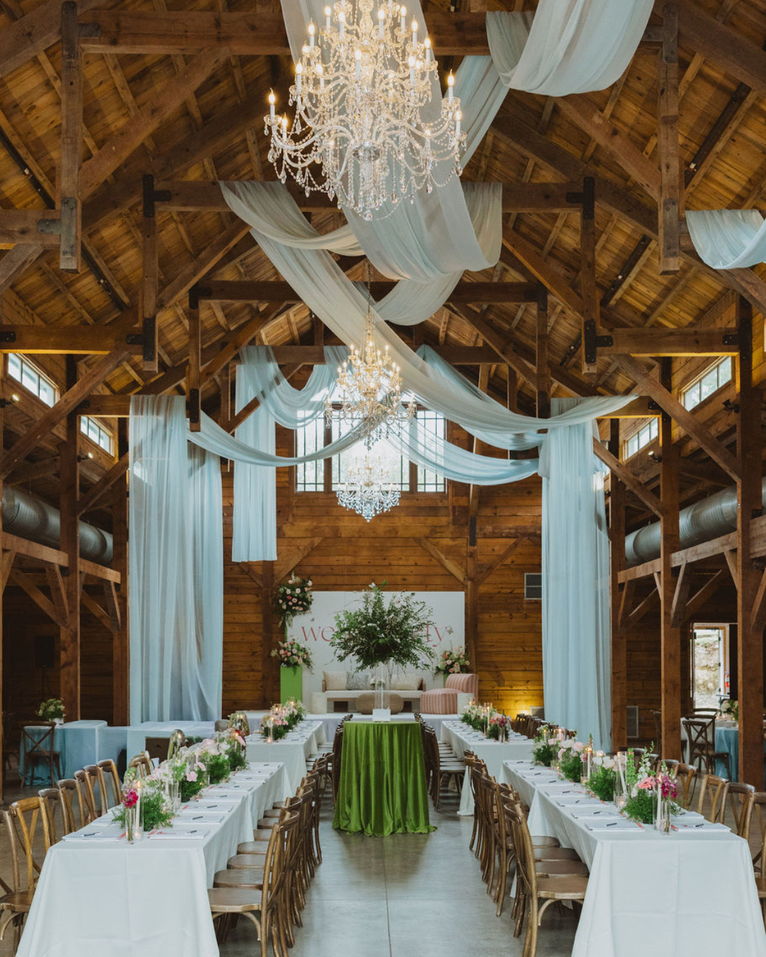 Rustic barn wedding reception with long white-clothed tables, chandeliers, and draped blue fabric