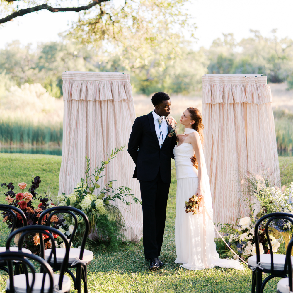 Wedding ceremony under floral arch, couple holding hands as officiant and guests look on.