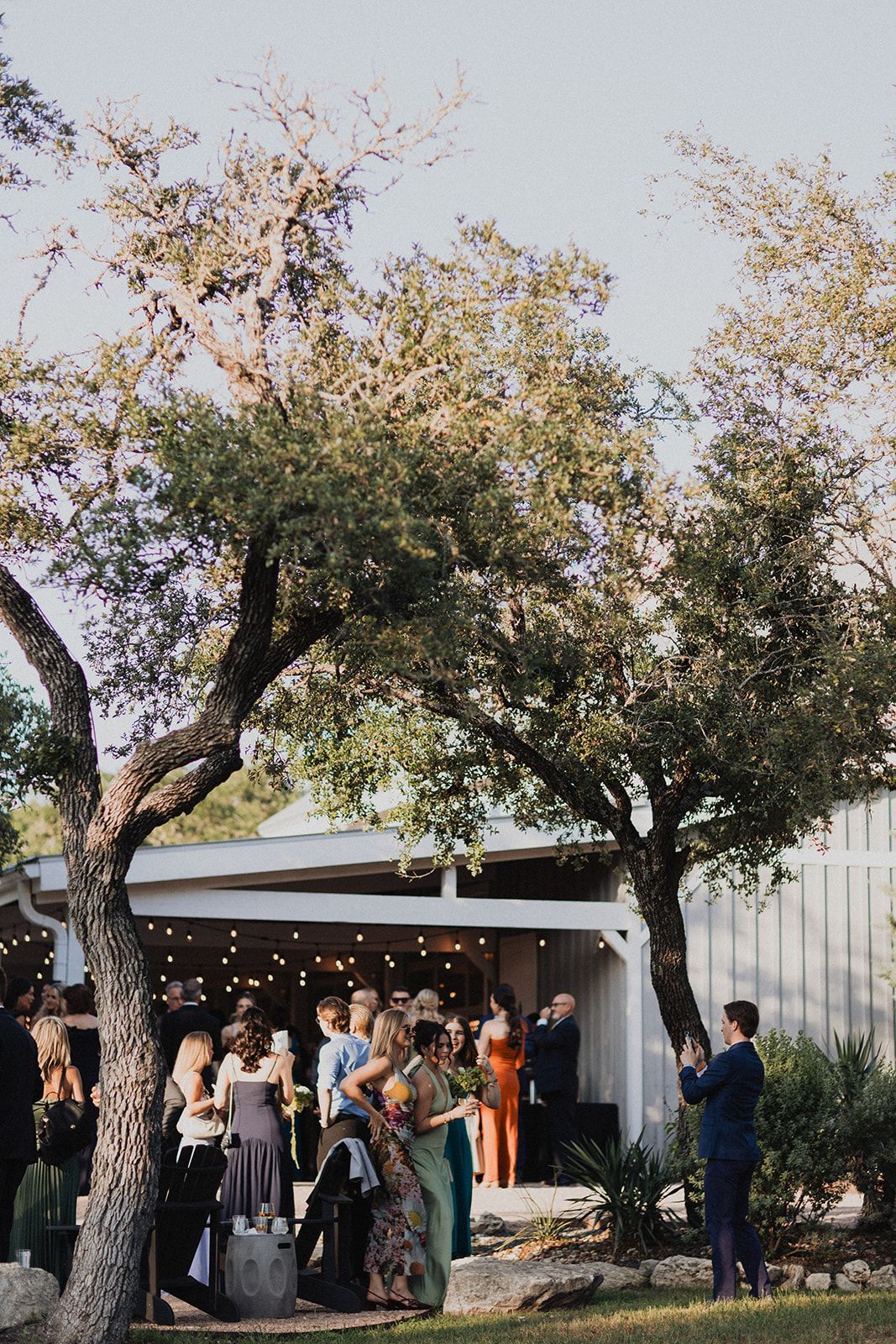 A group of people gathers for an outdoor evening event under trees near a lit building as one person takes a photo.