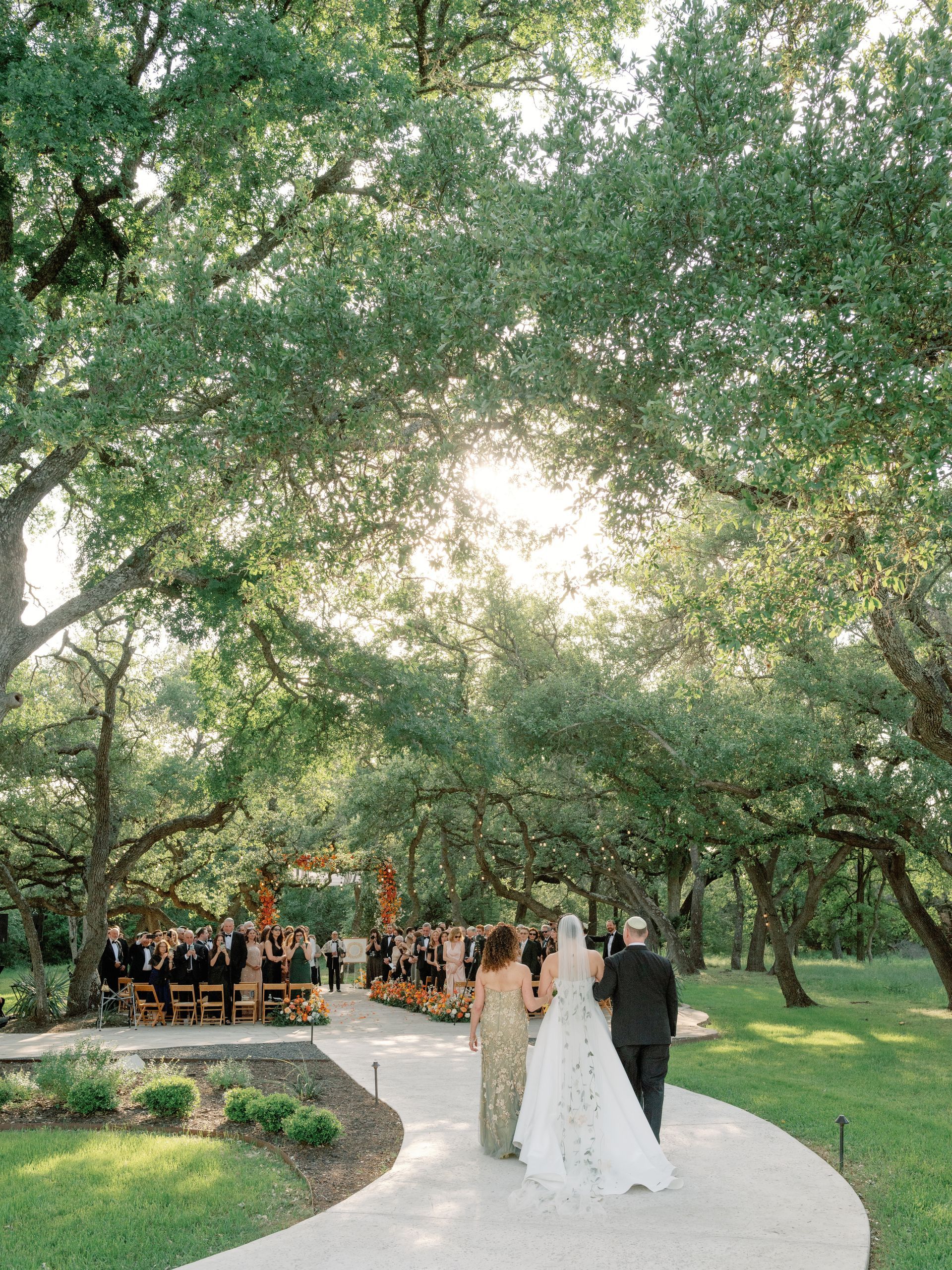 A bride in a white gown and a person in a dark suit walk down a path toward an outdoor wedding ceremony under large trees.