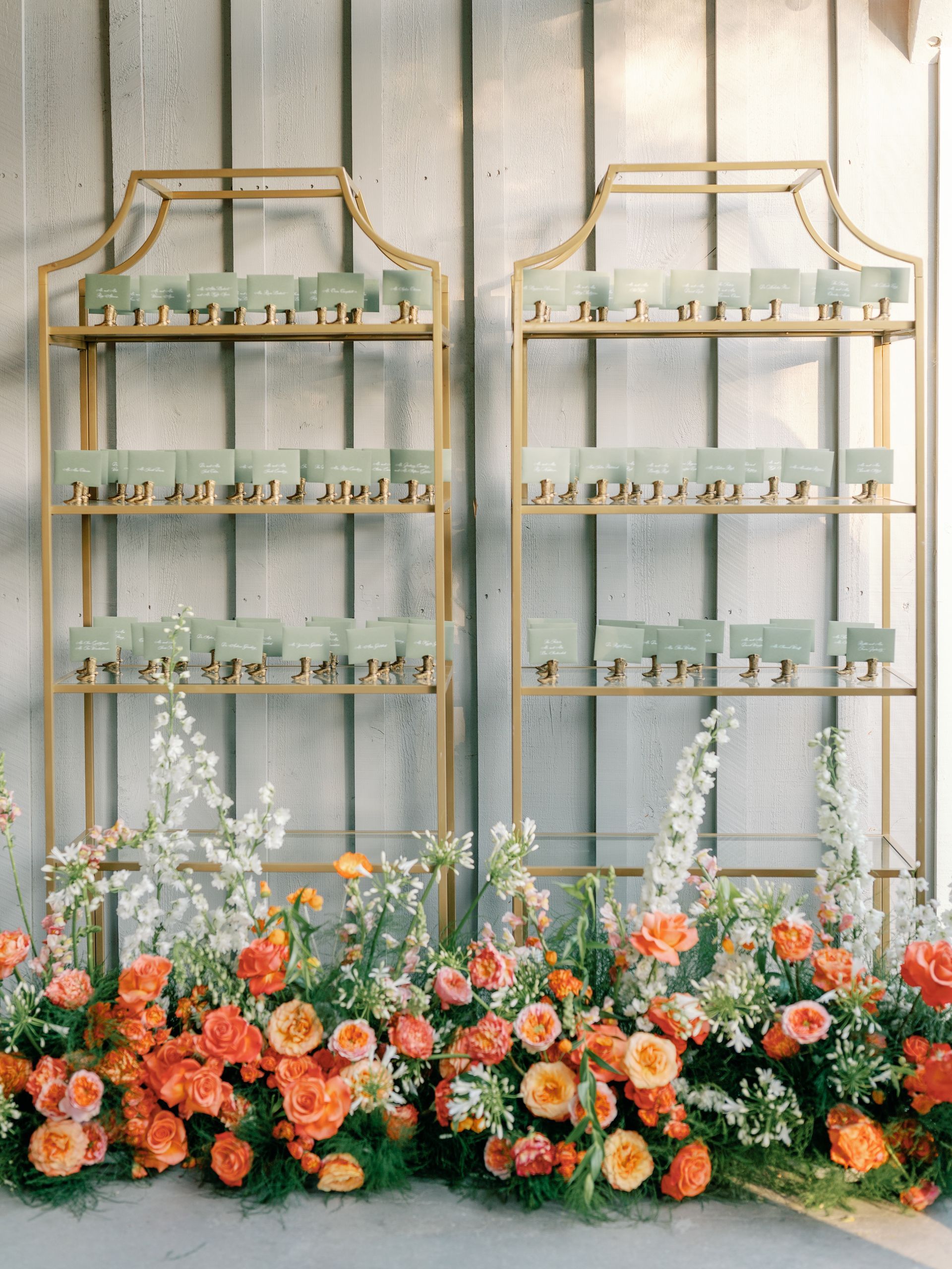 Two gold tiered shelves display green seating cards, fronted by a vibrant arrangement of orange and white flowers.