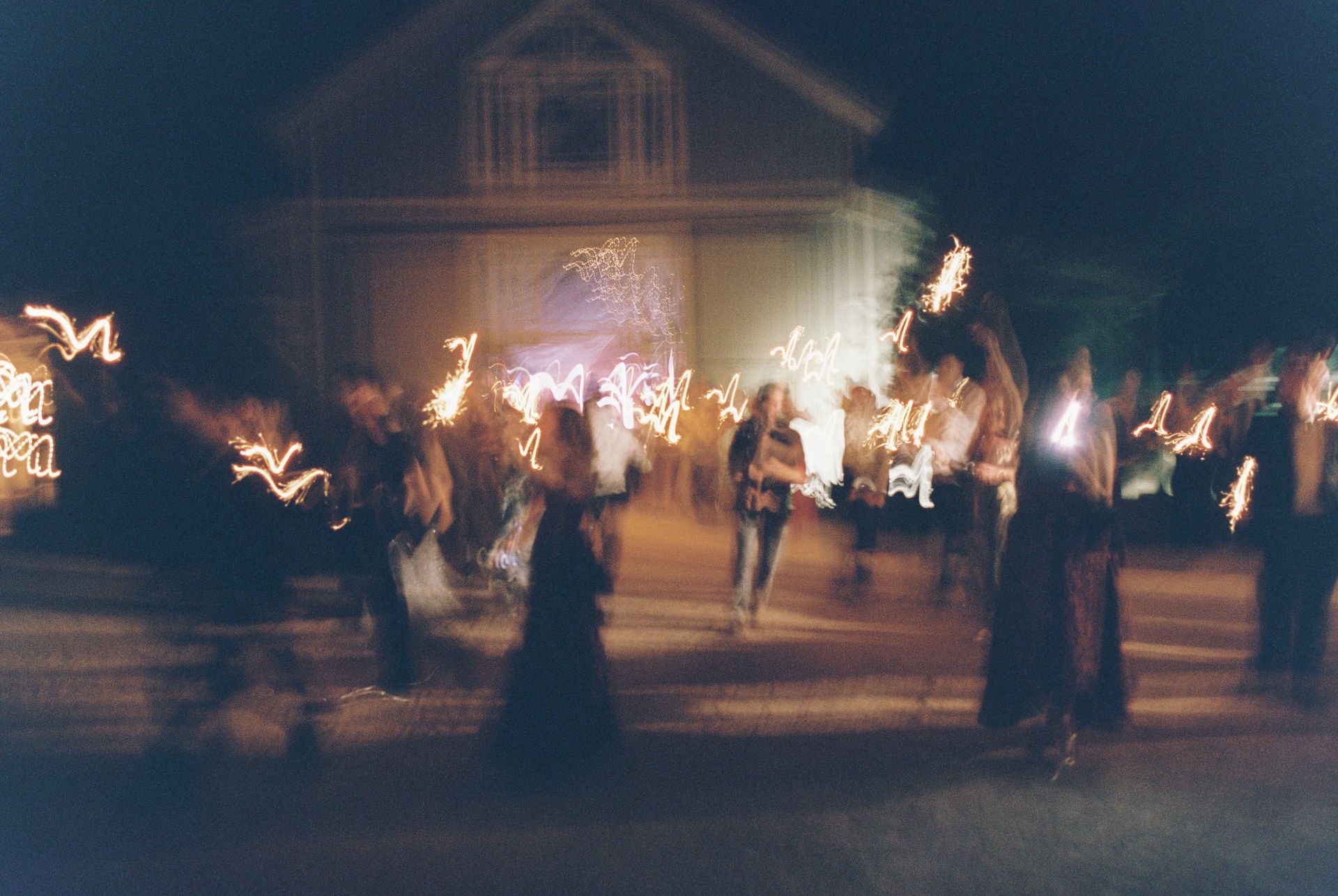 A blurred night scene shows people holding sparklers or light sources in front of a building.