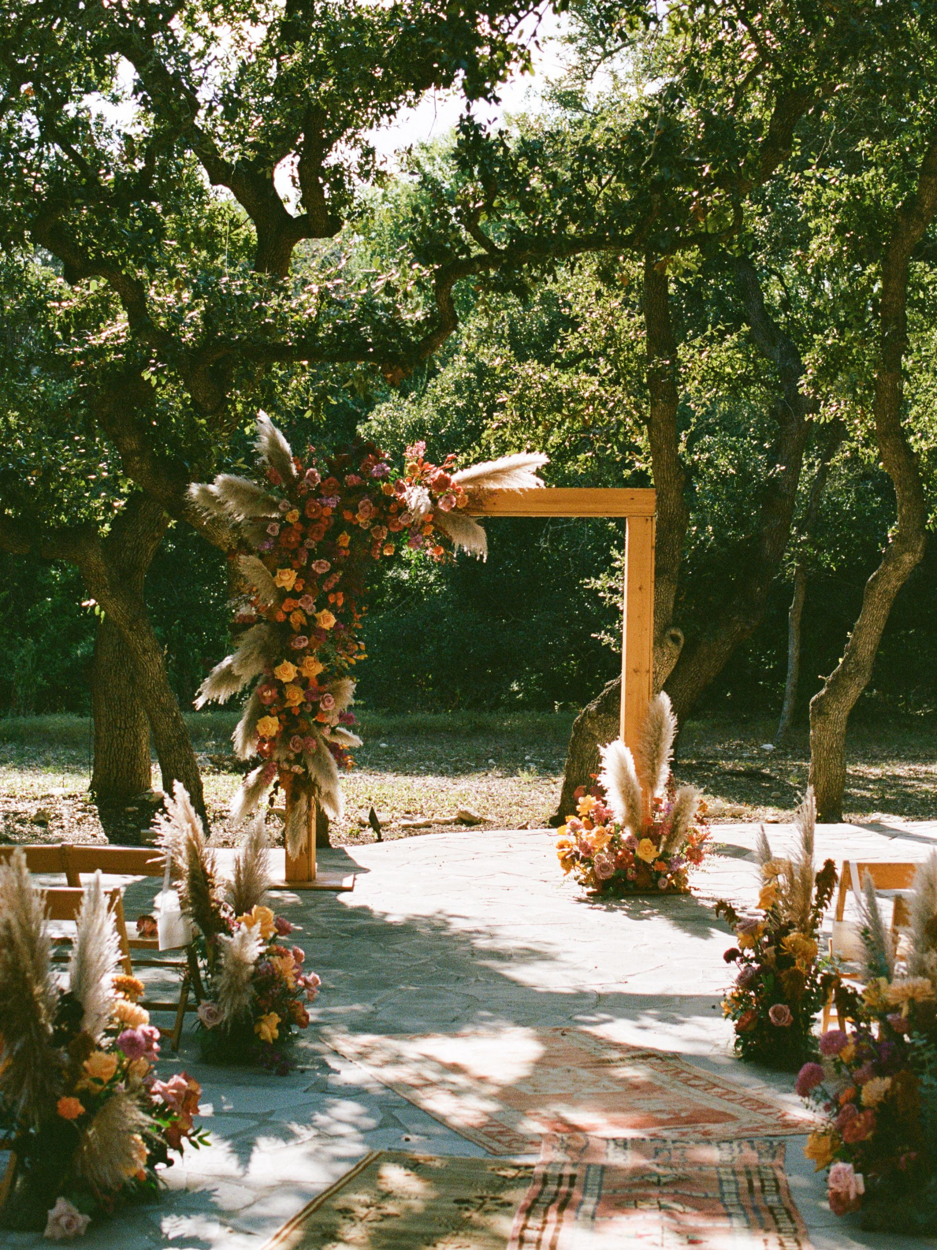 A rustic wedding ceremony altar featuring a wooden frame decorated with dried flowers and pampas grass in a forest setting.