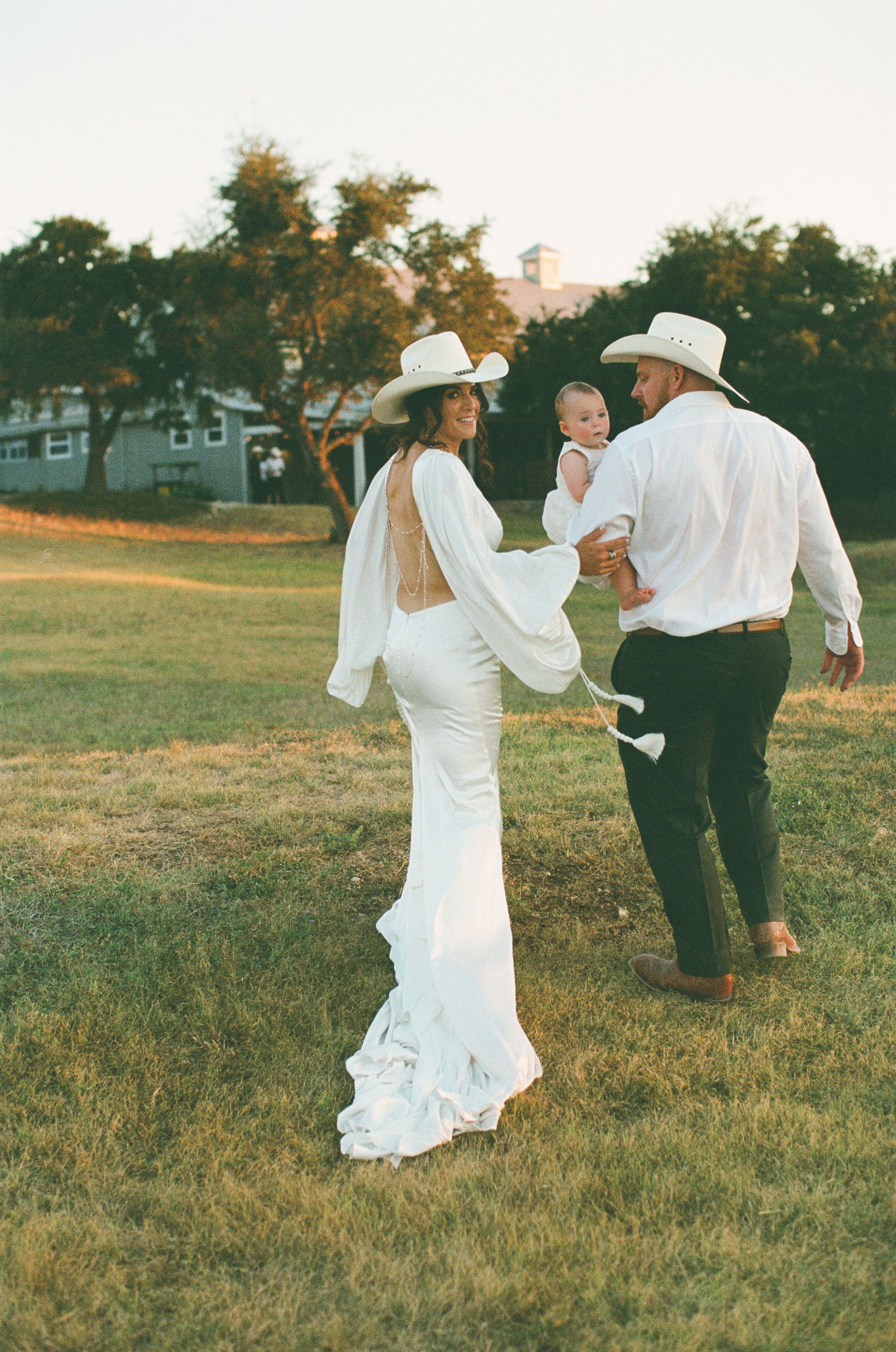 A couple in wedding attire and cowboy hats walking across a grassy field while carrying a baby.