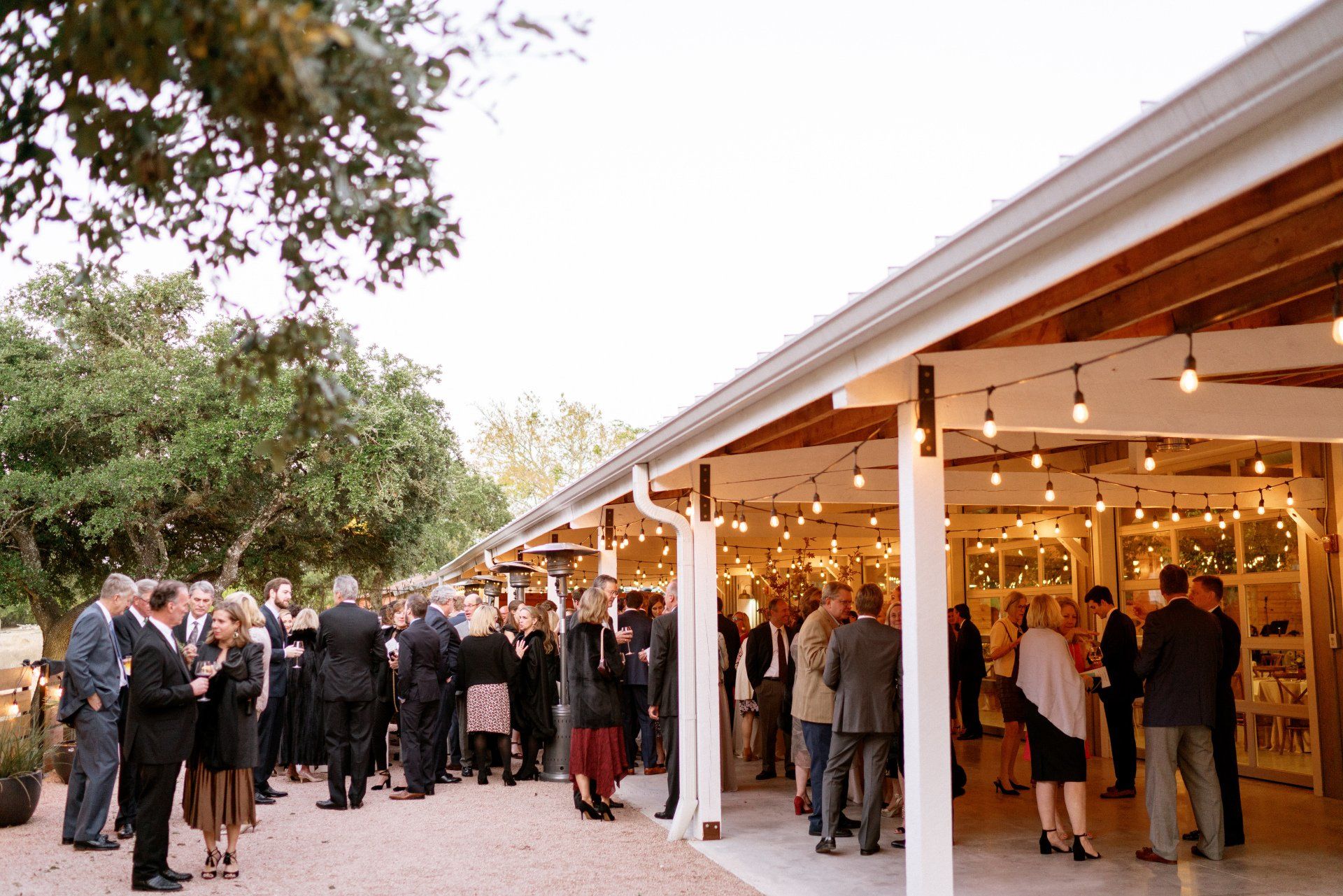 People in formal attire gather at an outdoor evening reception under a long, illuminated porch at a venue with trees.
